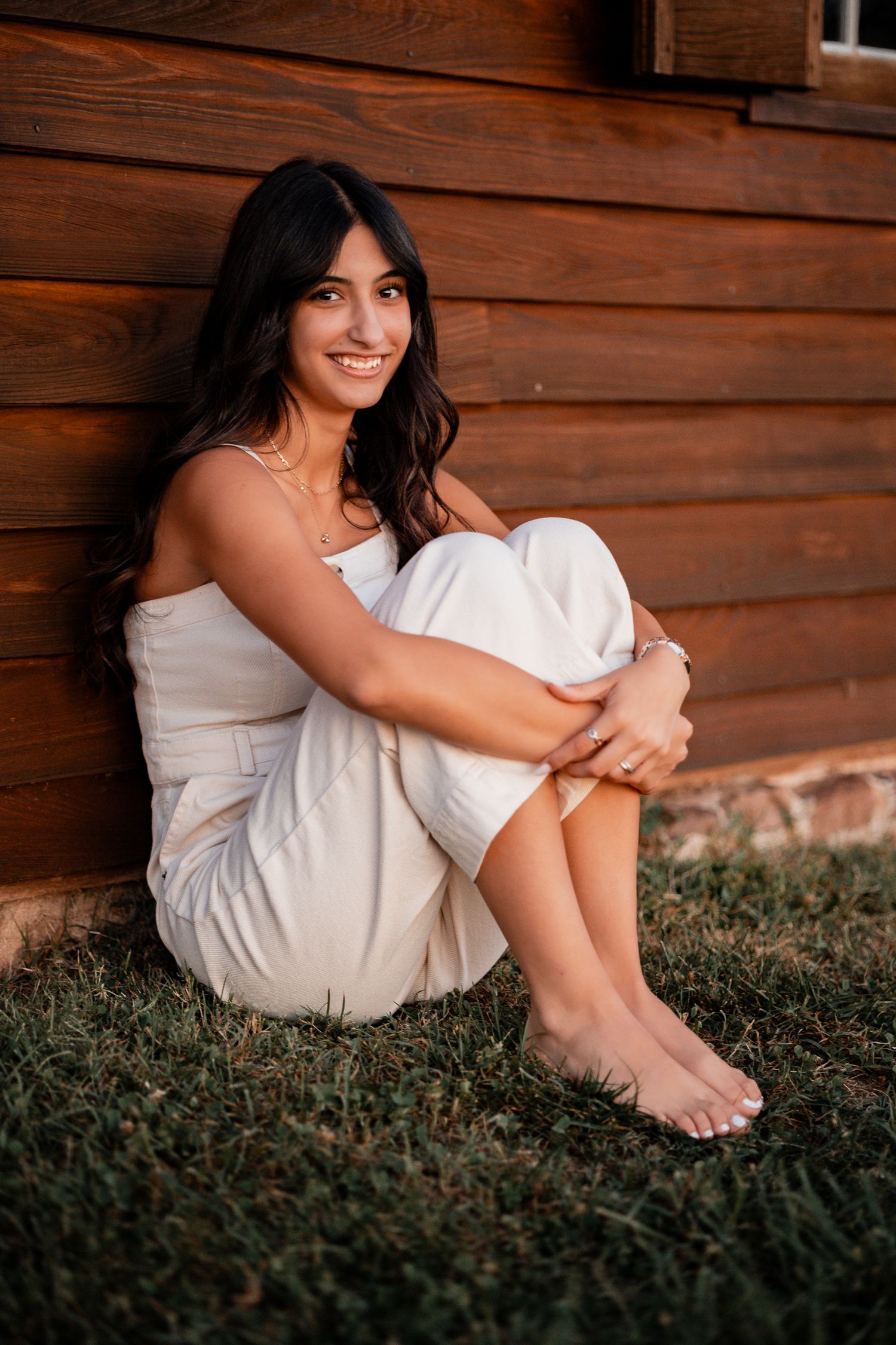 Candid portrait of someone in a white dress sitting by a rustic wooden wall at dusk.