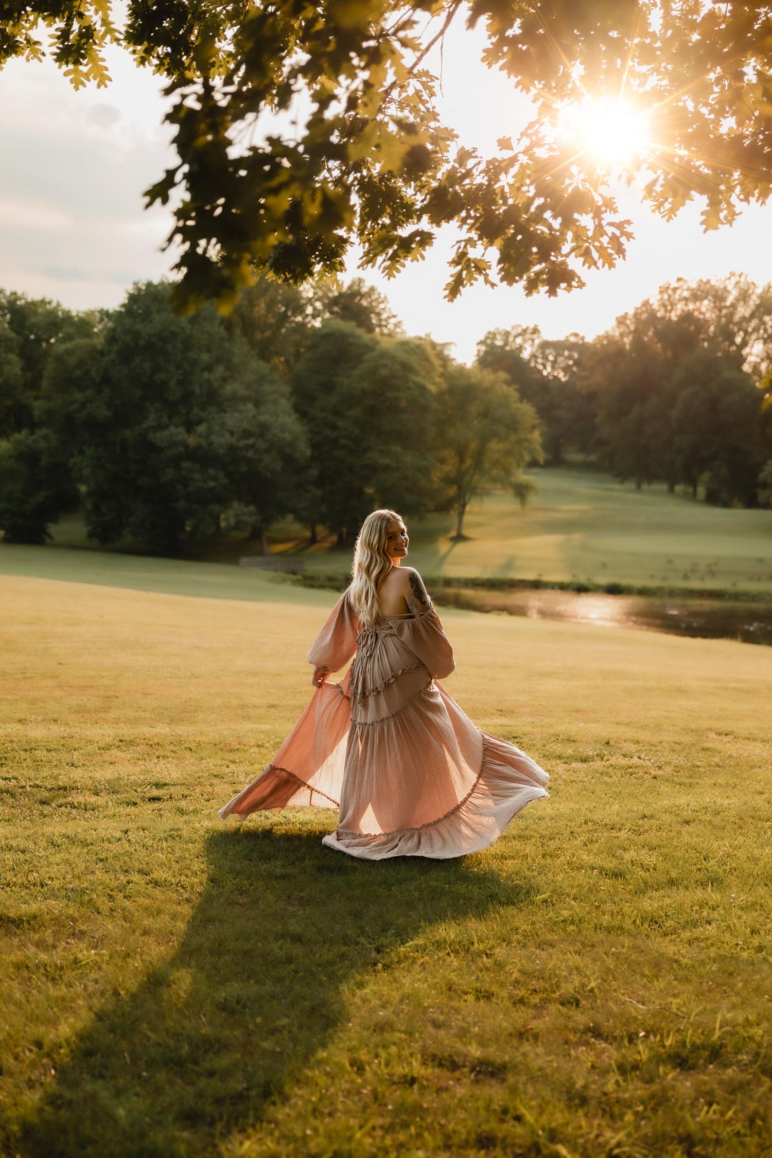 Woman in flowing pink gown walks through sunlit green park at golden hour, trees framing warm glowing sunset.