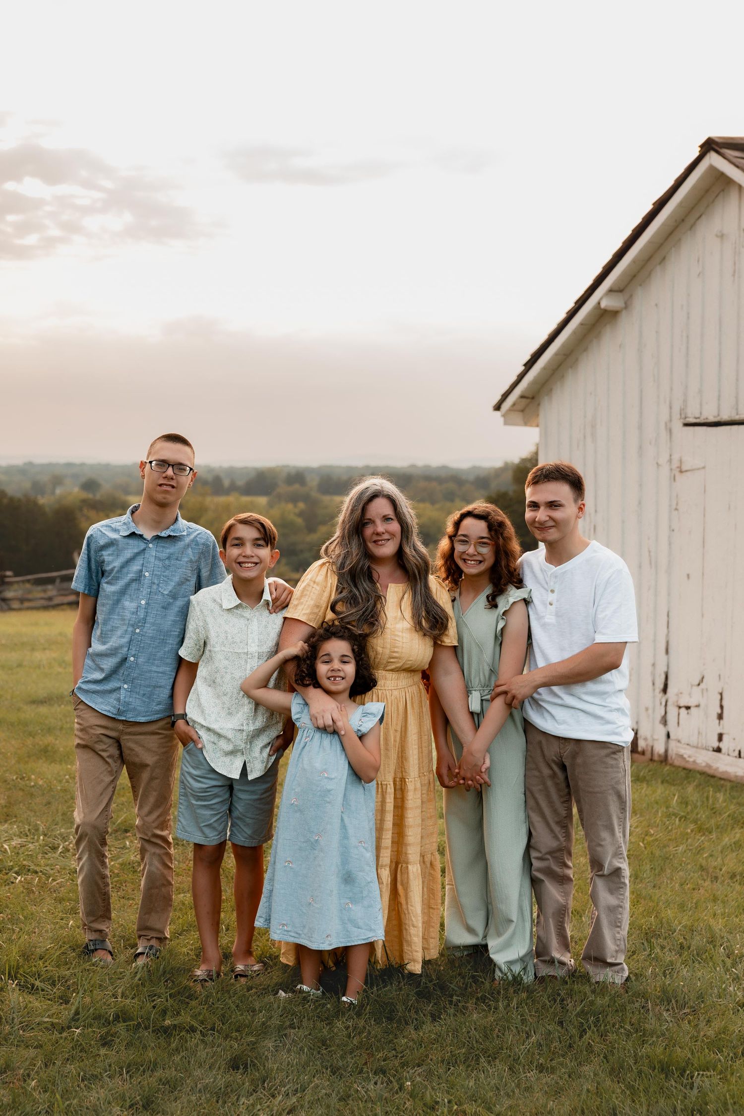 A family of six poses together in a field near a white barn at golden hour, wearing coordinated pastel outfits.