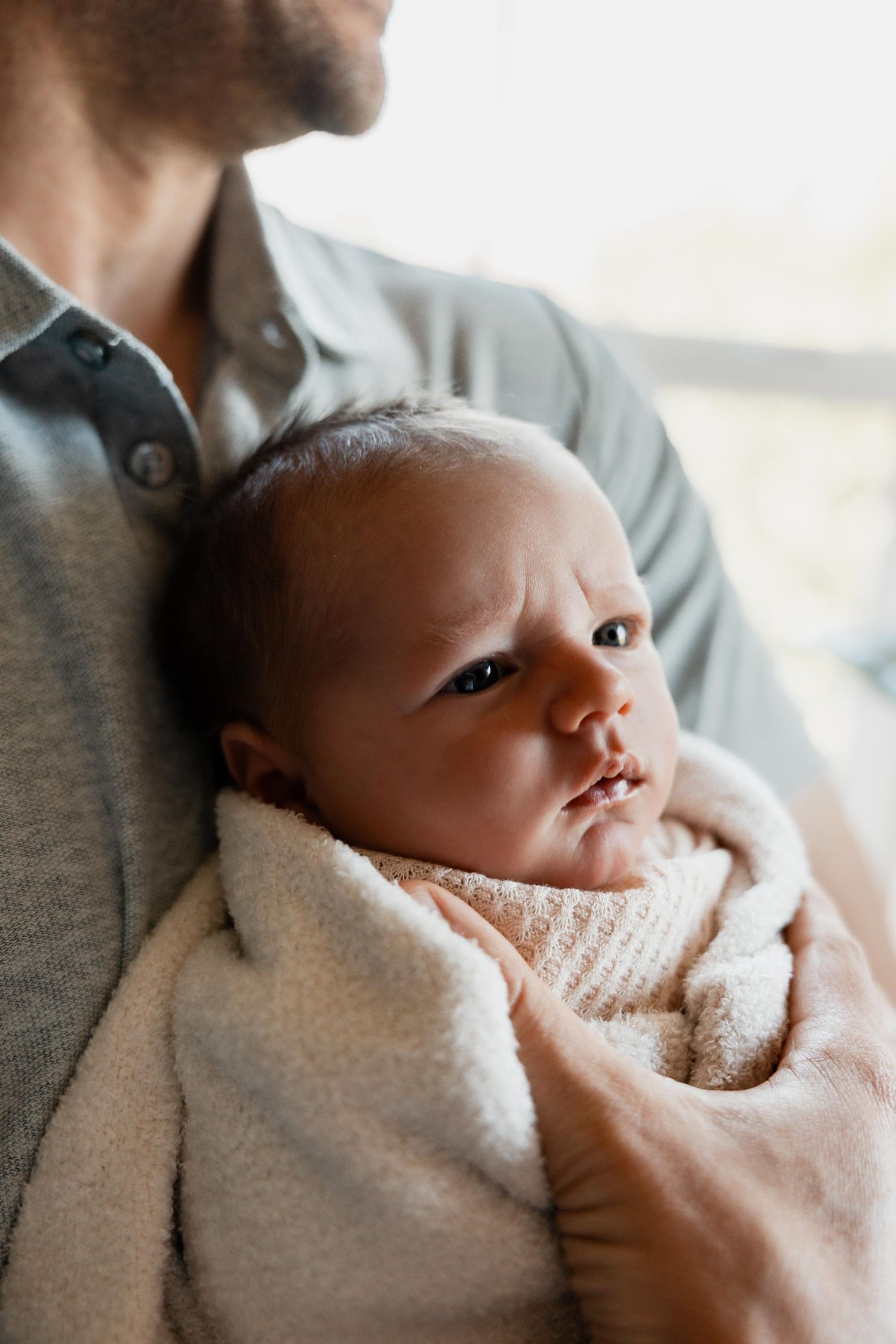 Close-up of alert newborn baby wrapped in soft blanket being held by father near a window.