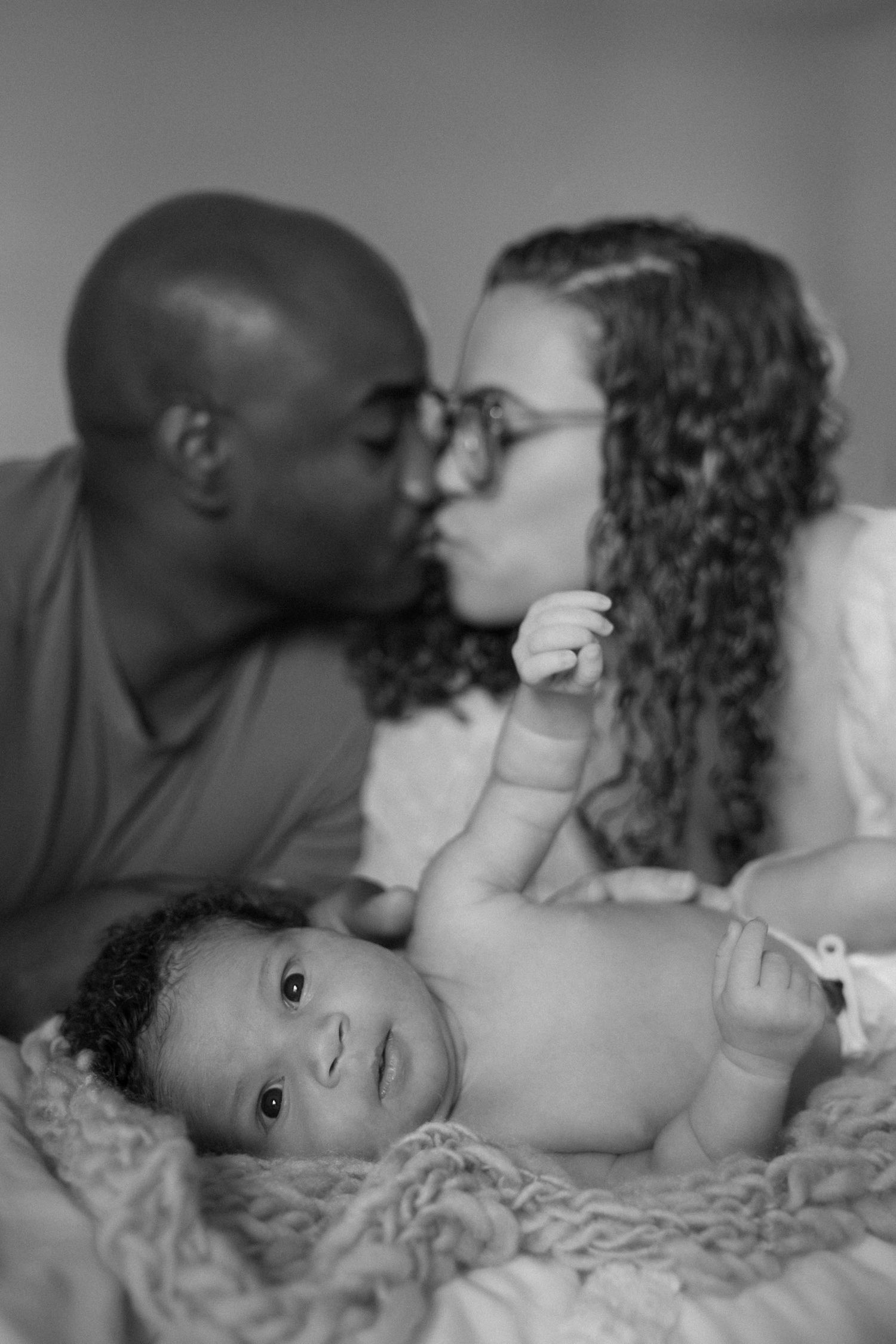 Black and white photo of interracial couple kissing behind their newborn baby lying on a knit blanket.
