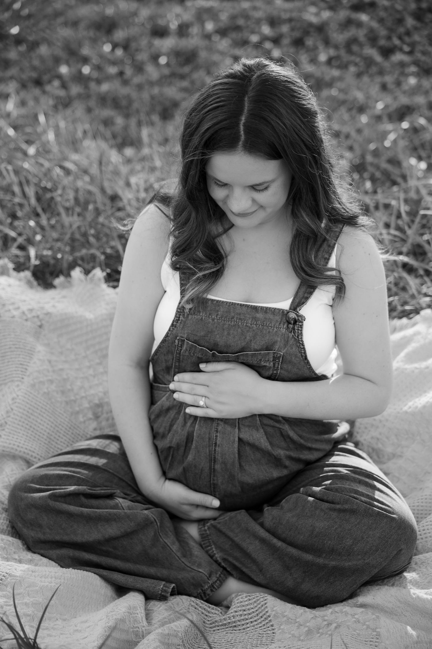 Black and white photo of pregnant woman in denim overalls sitting cross-legged, cradling her baby bump outdoors.