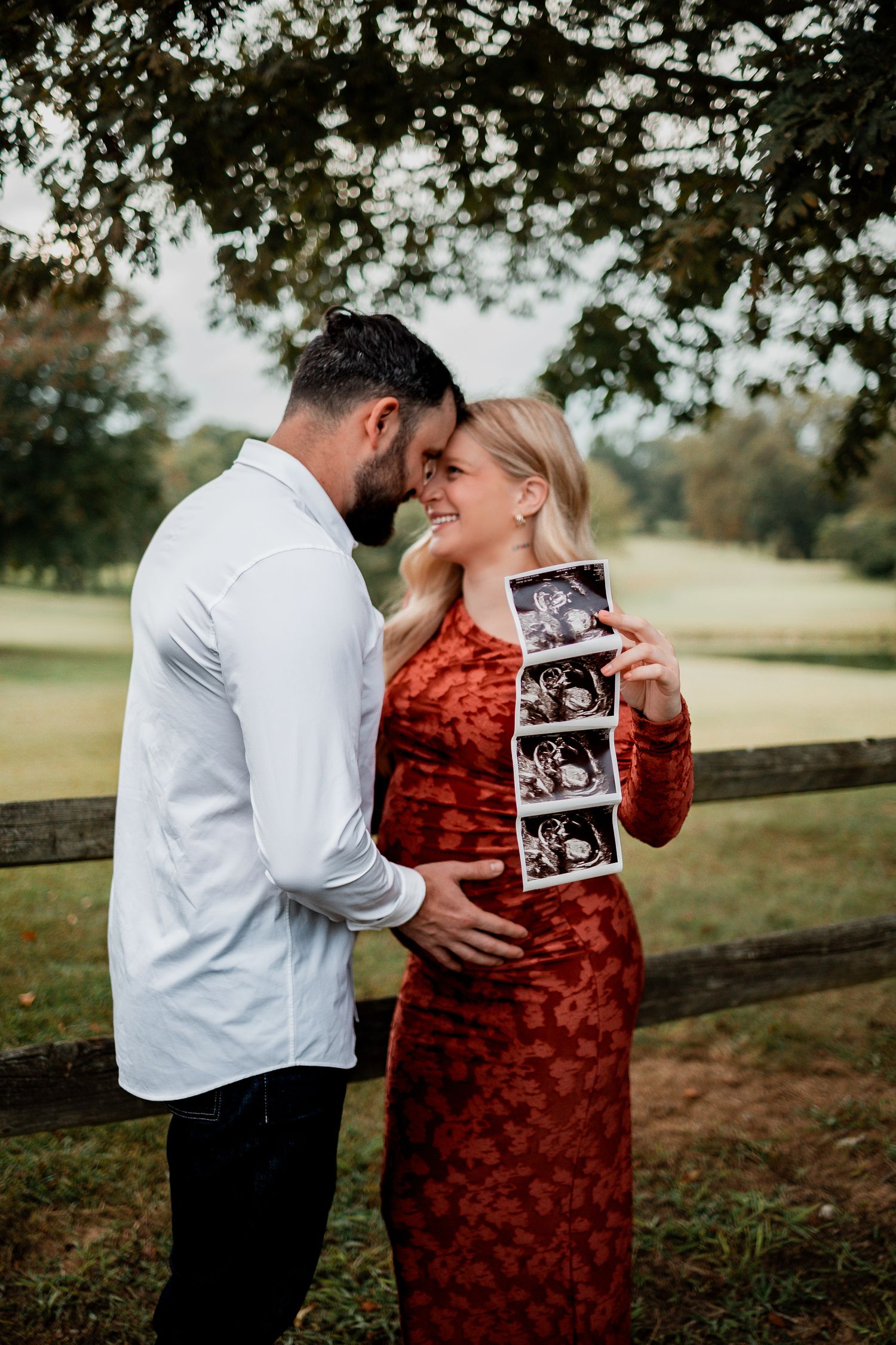A couple embraces outdoors while holding an ultrasound photo, sharing a tender moment in a rustic setting.