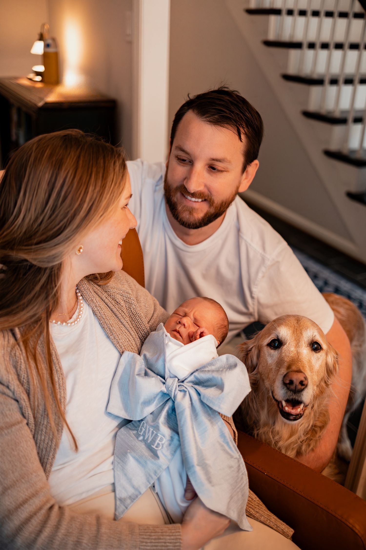 A family sits together on stairs with their golden retriever and newborn baby wrapped in blue blanket.