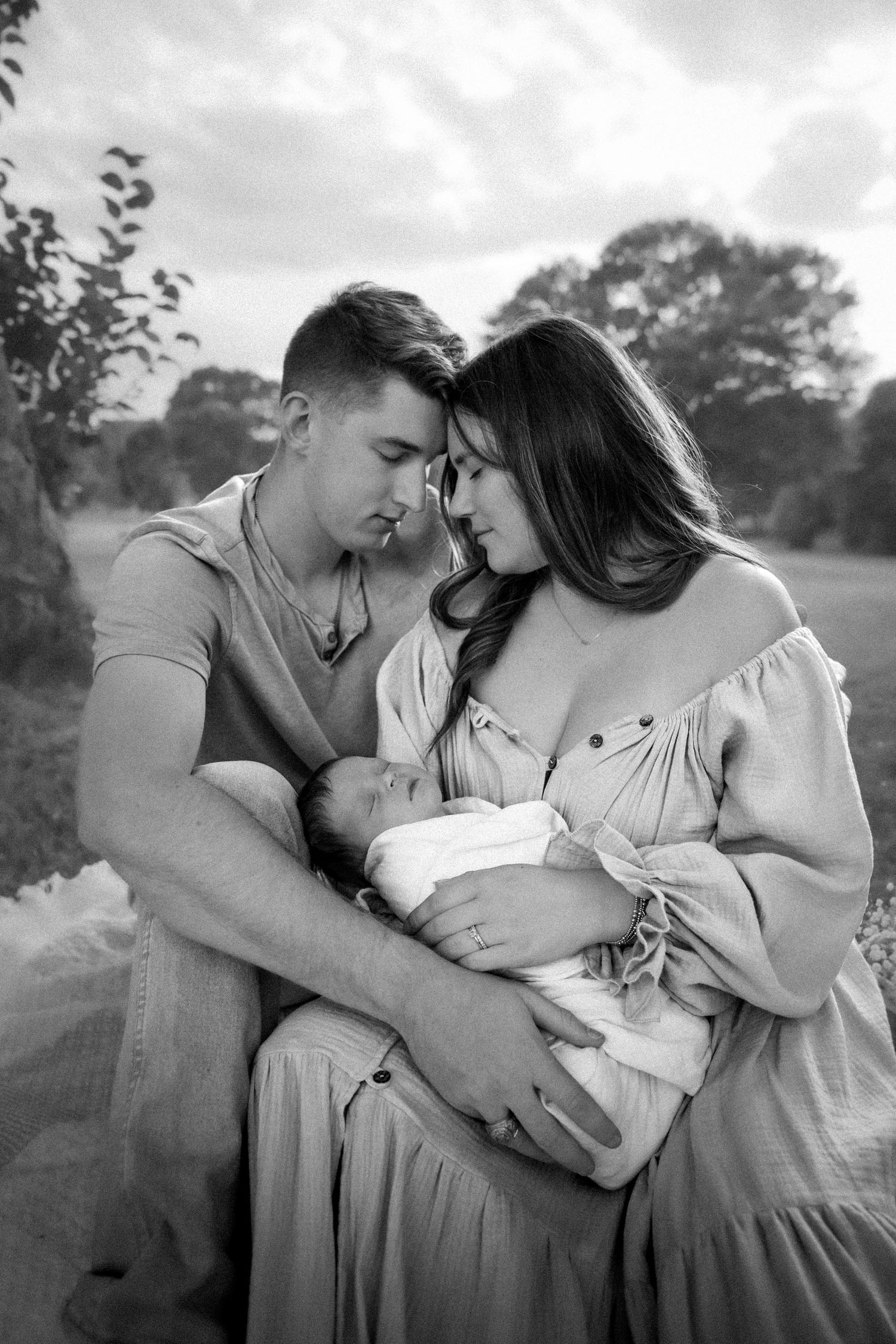 Black and white photo of a couple tenderly holding their newborn baby outdoors, foreheads touching in an intimate moment.