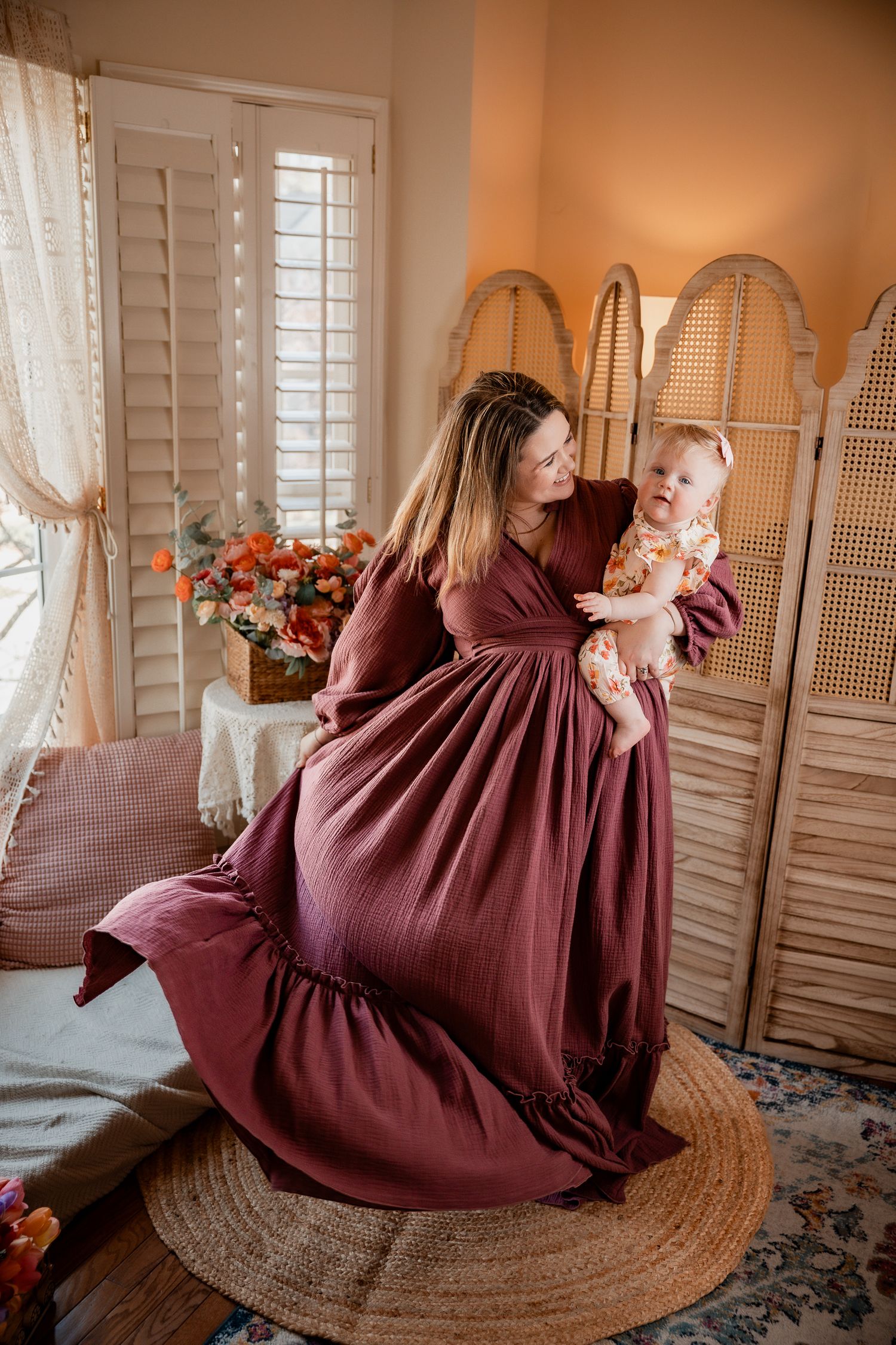 A mother in a burgundy maxi dress twirls while holding her baby in a floral outfit in a warmly decorated room.