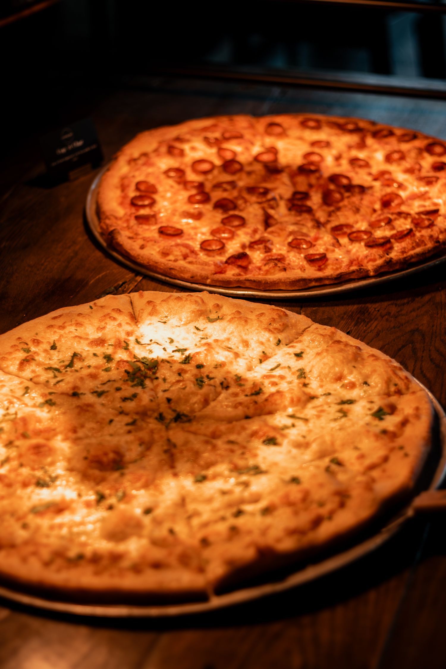 Two pizzas on wooden tables in dim lighting, one with pepperoni and one with herbs and cheese.