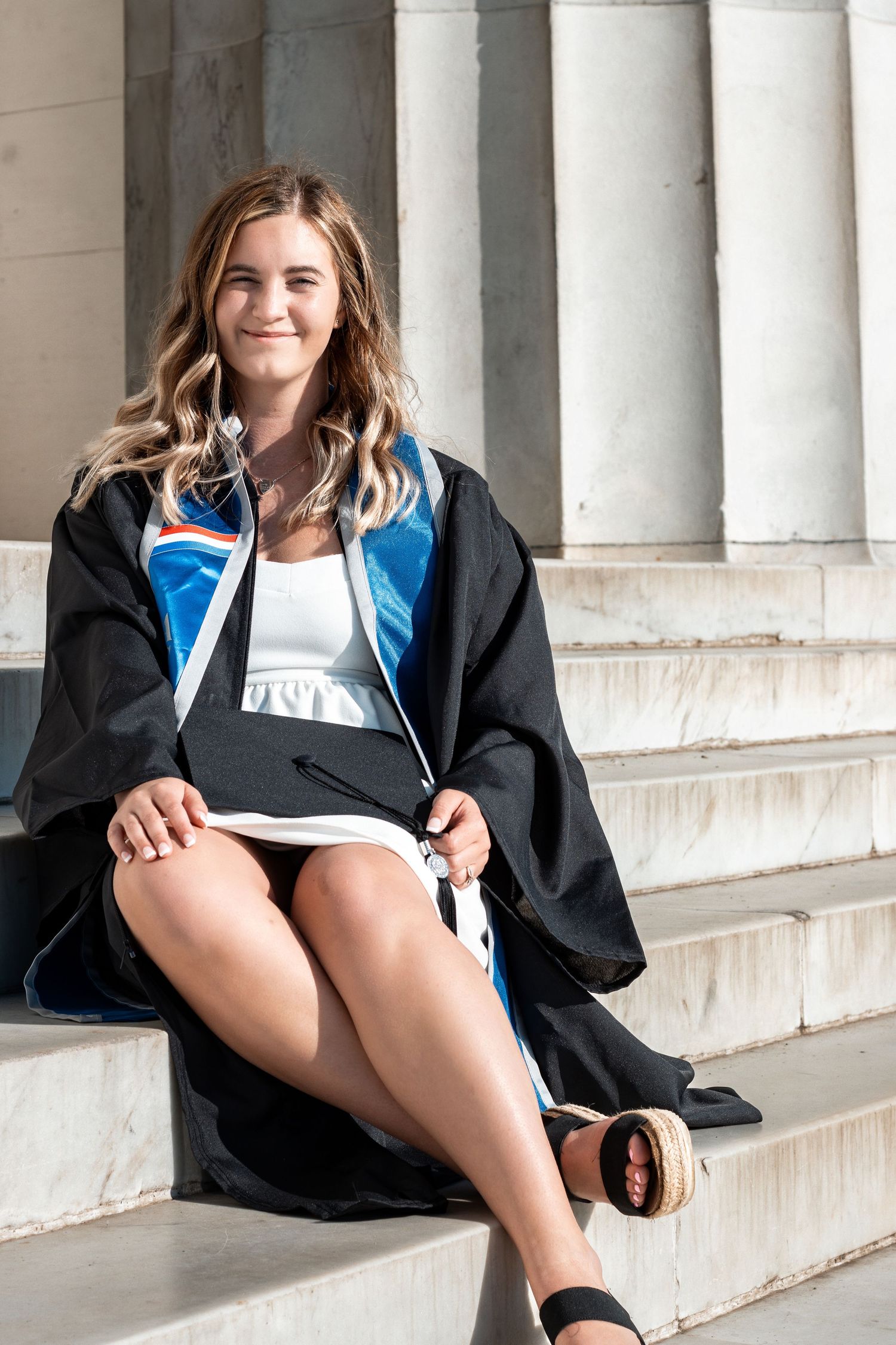Graduate seated on stone steps wearing black graduation gown and blue stole in natural lighting.