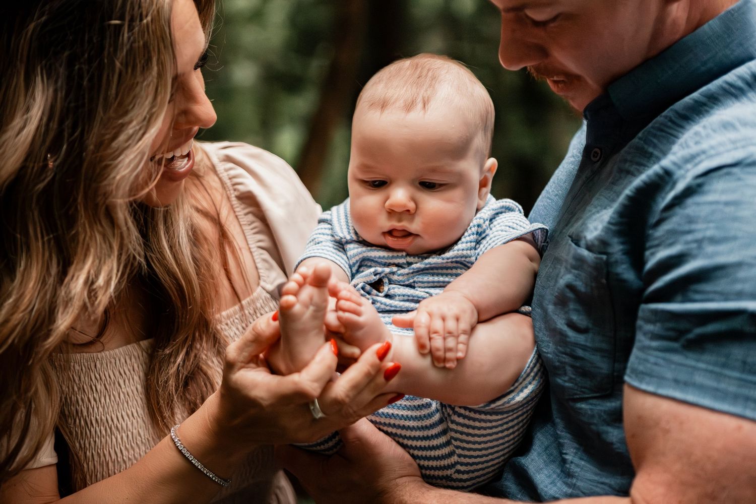 Baby in a striped outfit reaches out while being held by parents in an intimate family moment outdoors.