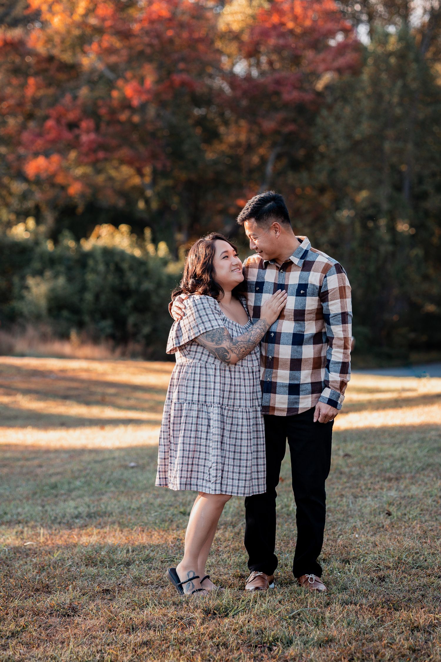 A couple in matching plaid outfits share a tender moment in a park with vibrant autumn foliage in the background.