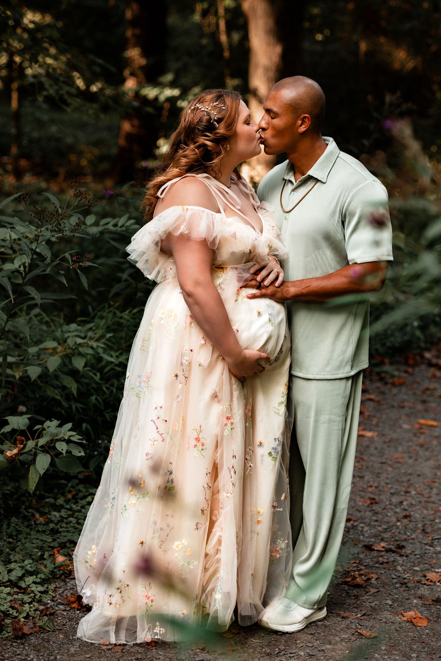 A couple shares romantic moments in a wooded outdoor setting wearing light colored formal attire.