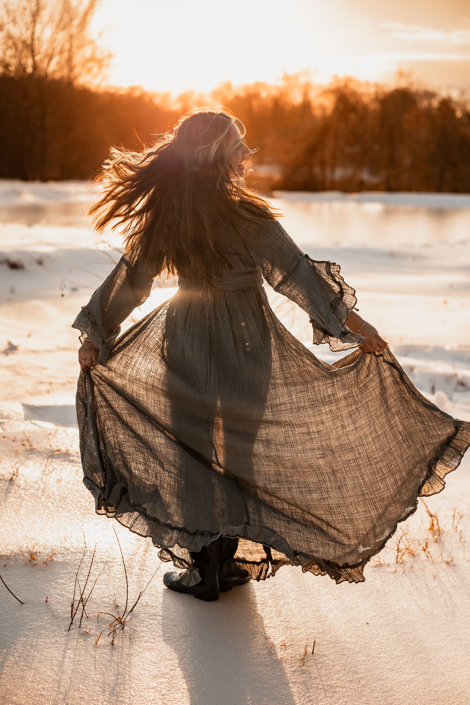 Woman in flowing gray dress dancing in snowy field at golden sunset, hair blowing in wind.