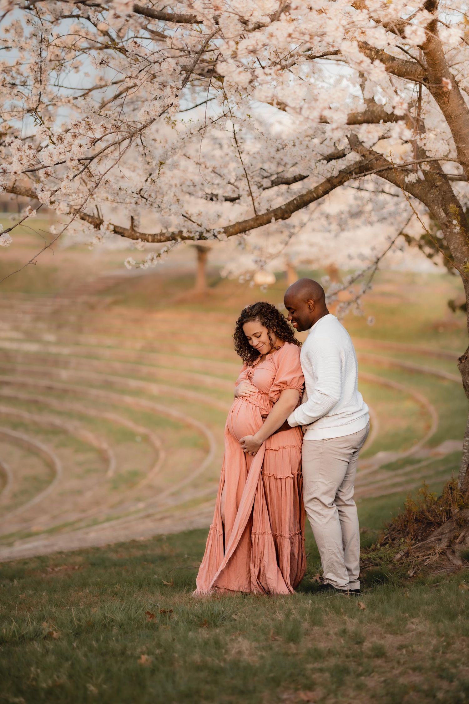 Pregnant couple embracing under cherry blossom tree, woman in pink maxi dress, man in white sweater, spring maternity photo.