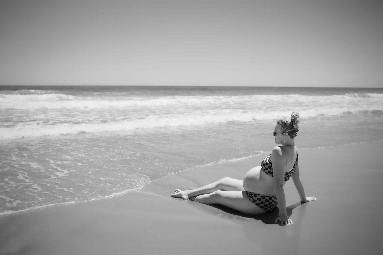 Black and white photo of a woman in a bikini sitting on a sandy beach with ocean waves in the background.