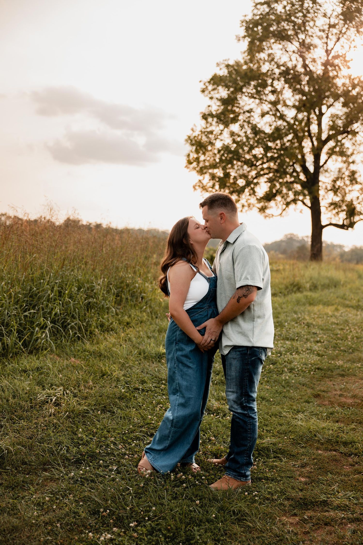 Couple sharing a tender kiss in a golden sunset field, woman in denim overalls, man in light shirt and jeans.