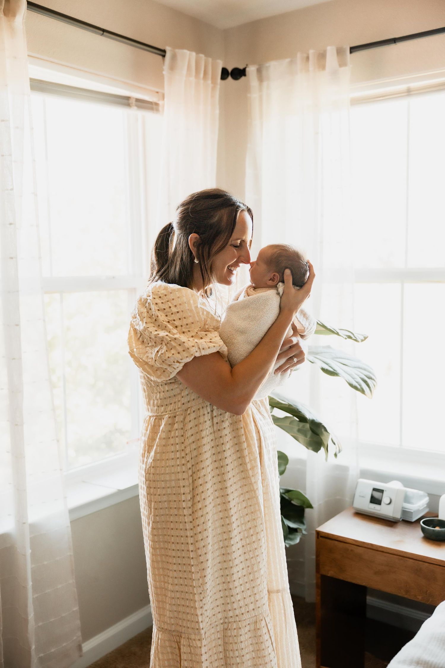 Mother in cream lace dress tenderly holding newborn baby near bright window in nursery room.