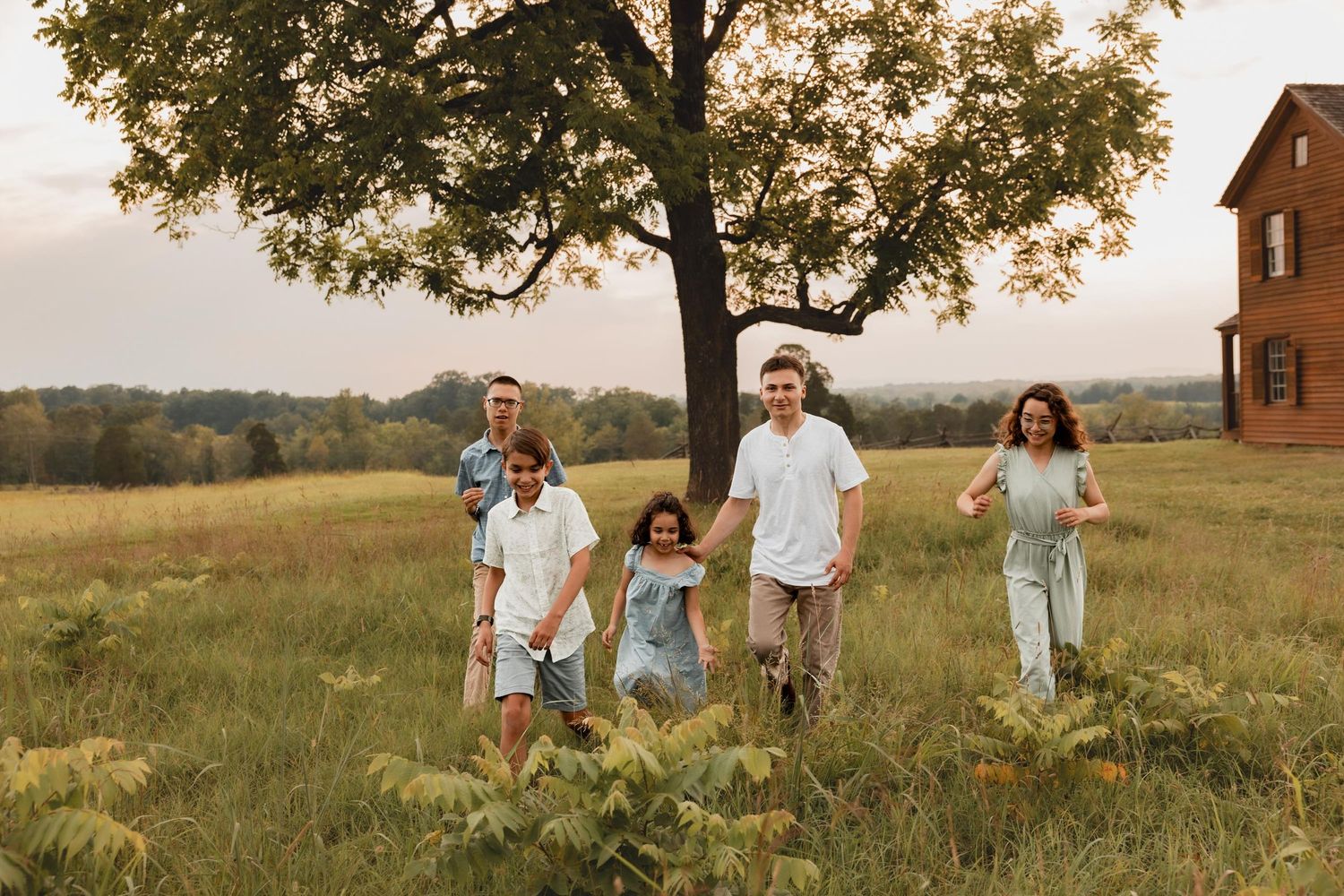 Family of five walking through a lush green field at sunset near a large tree and red barn.
