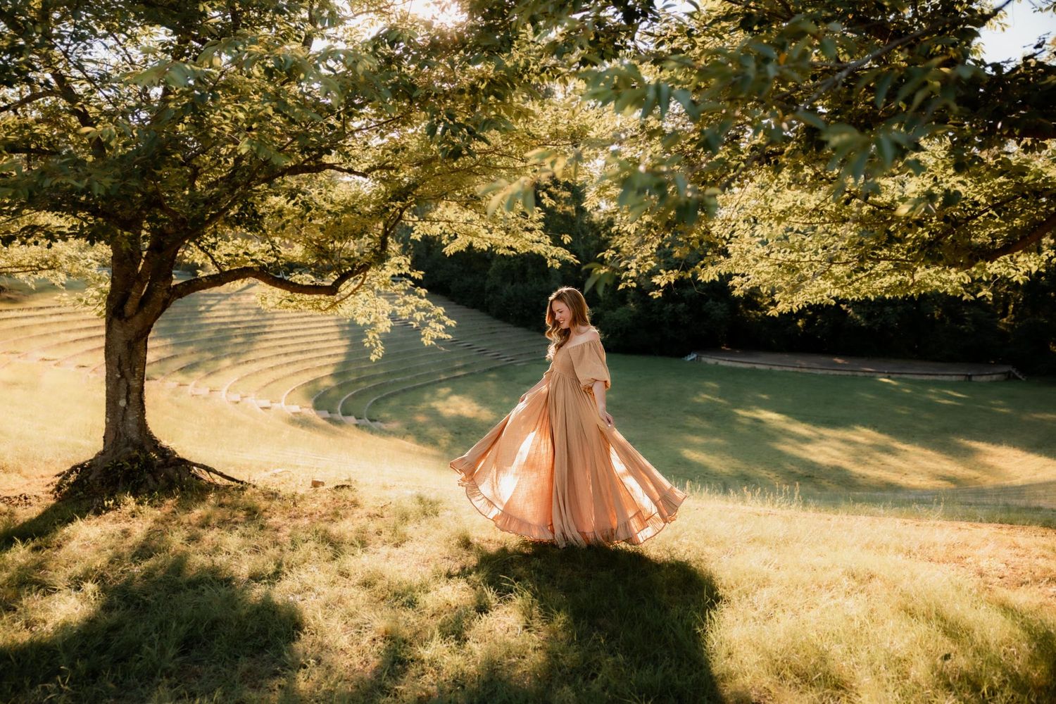 Woman in flowing peach dress twirling under a large tree in a sunlit green park.