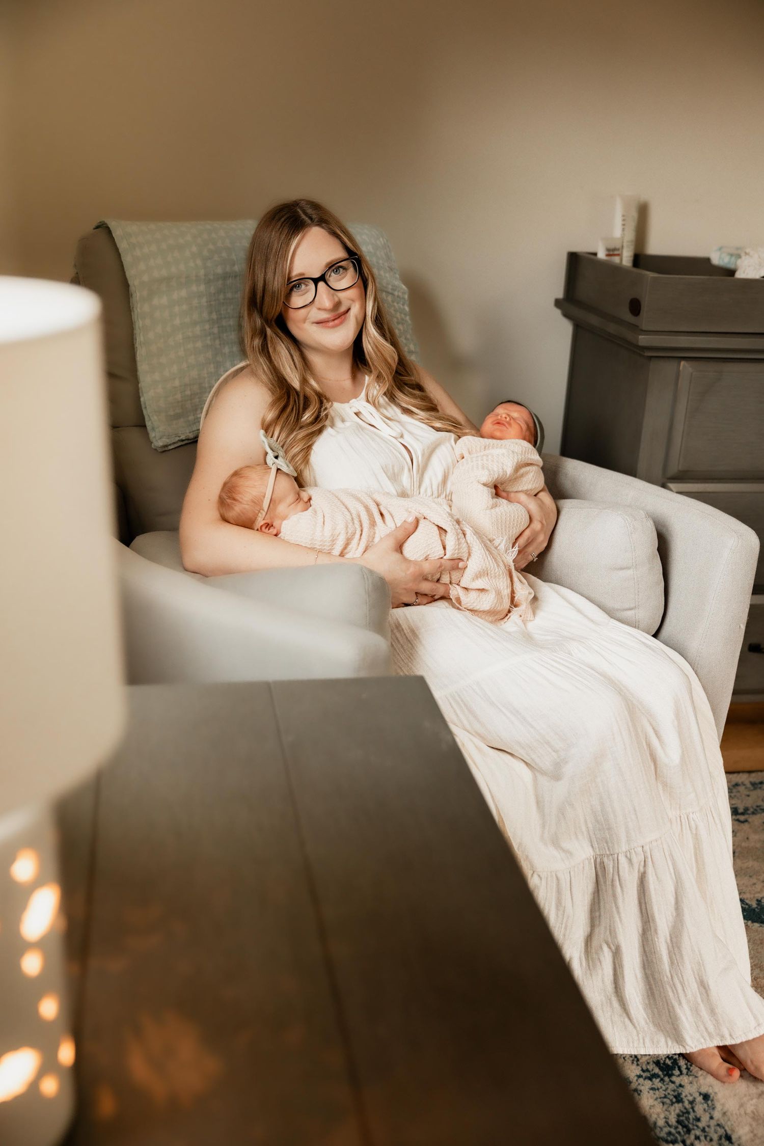 A smiling mother in a white dress and glasses sits in a nursery chair holding two newborn babies.
