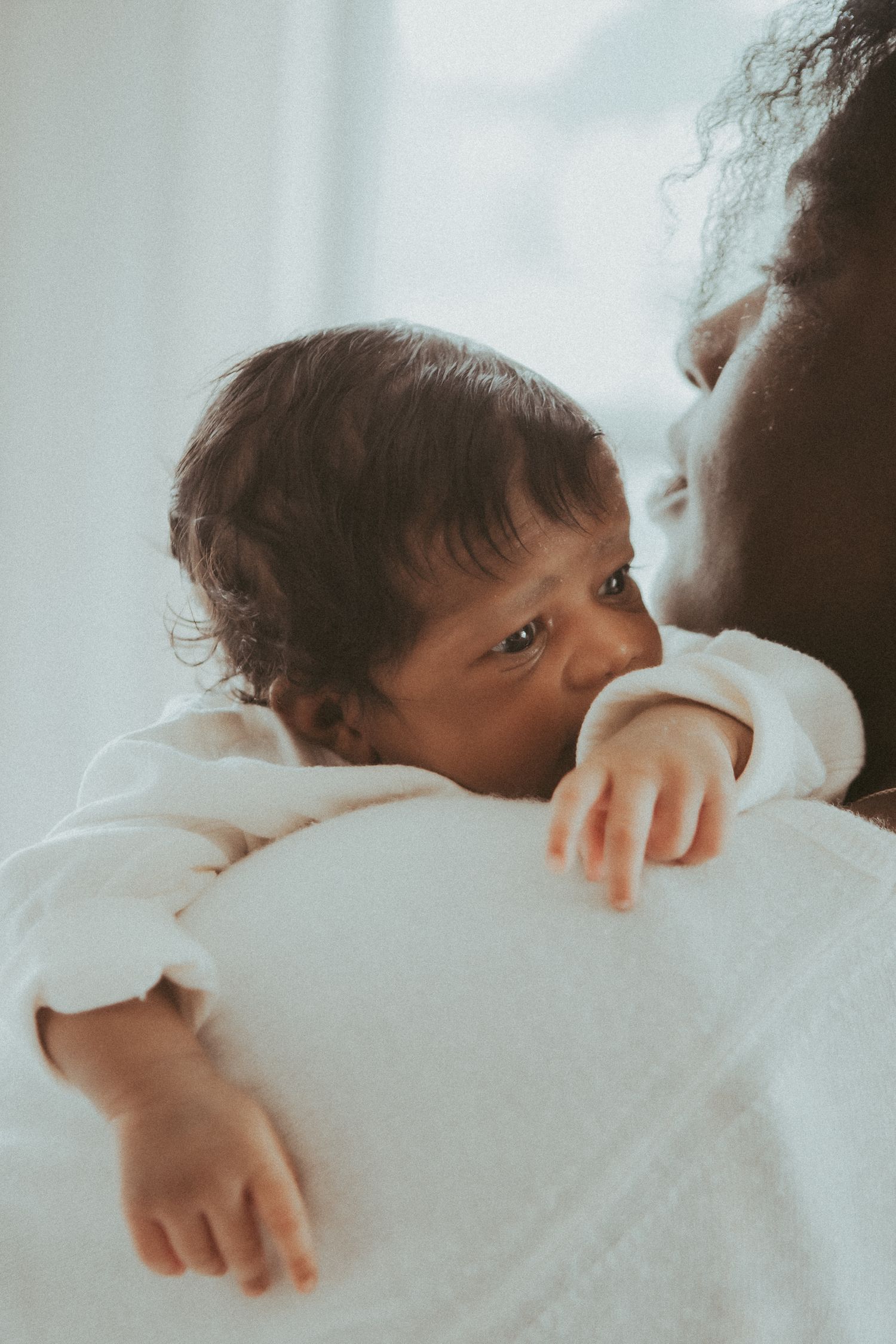A tender moment showing a baby resting on someone's shoulder wearing a white garment against a soft light background.