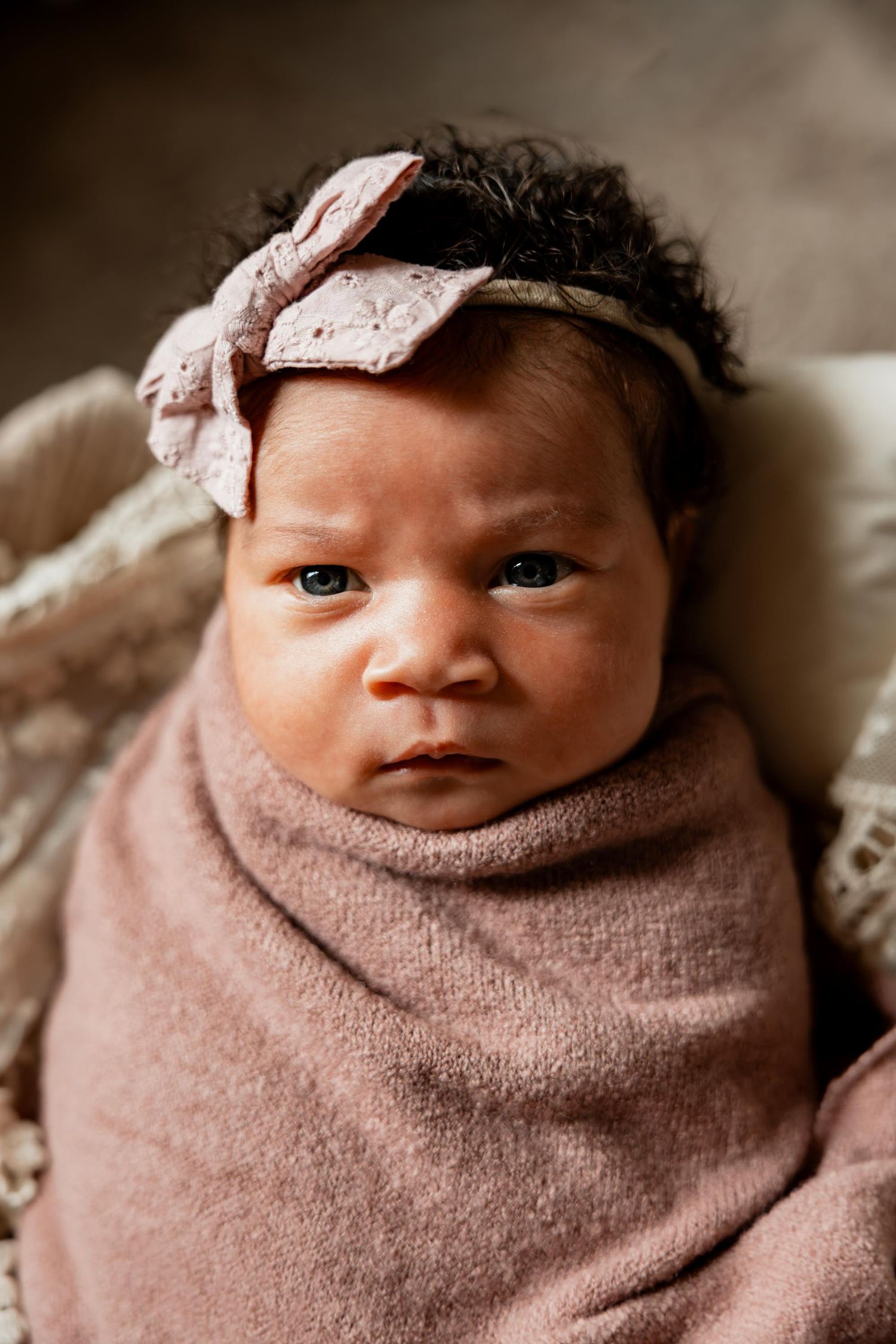 Newborn baby girl swaddled in pink wrap with bow headband, alert dark eyes, curly black hair, studio portrait.