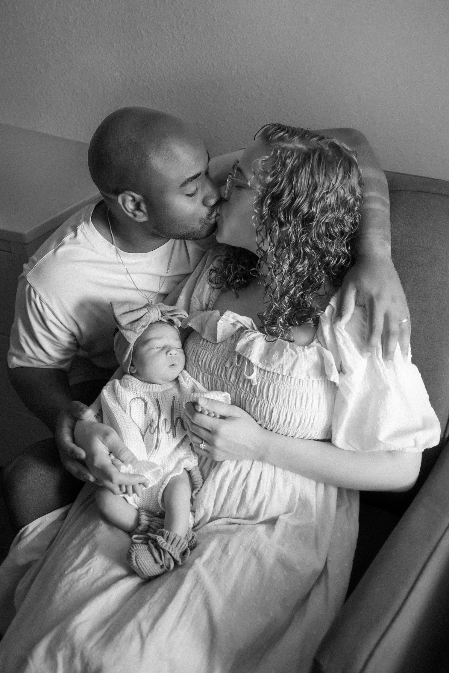 Black and white photo of a couple kissing while holding their newborn baby, a tender family portrait moment.
