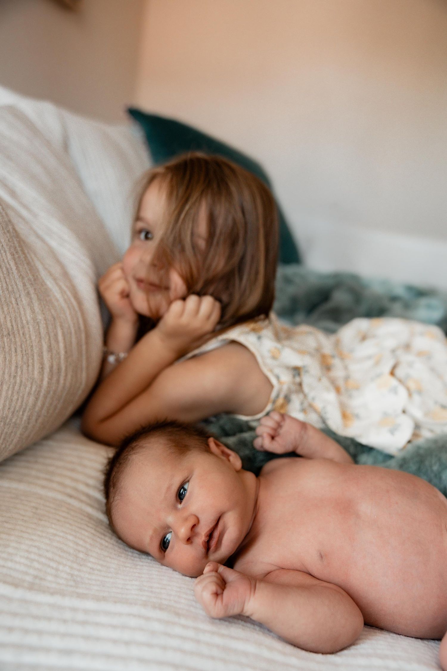 Newborn baby lying on bed with older toddler sibling smiling behind, cozy home setting.