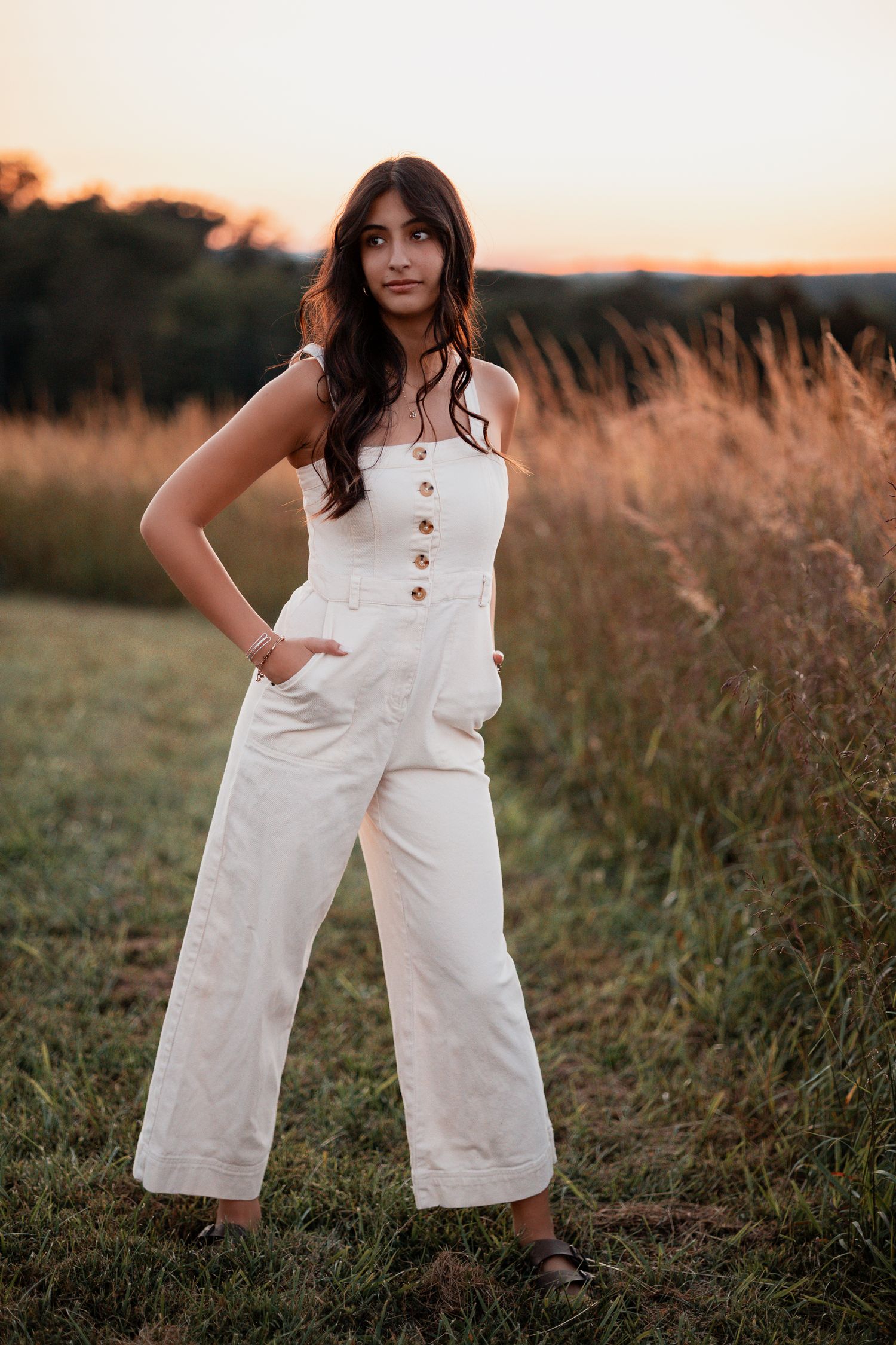 Person wearing white jumpsuit poses casually in field during sunset.