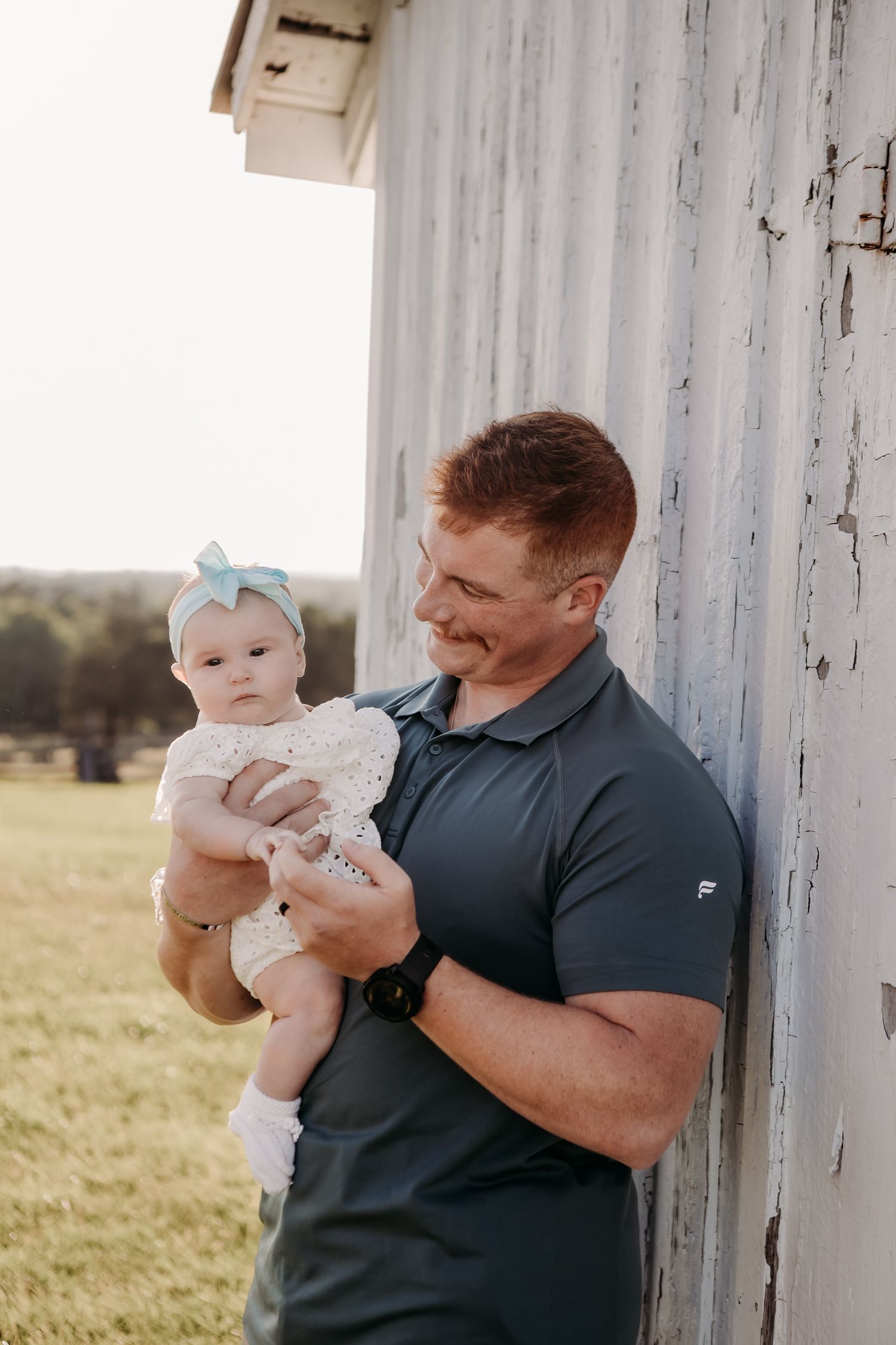 A baby wearing a bow and white dress is held outdoors next to a rustic white barn wall.