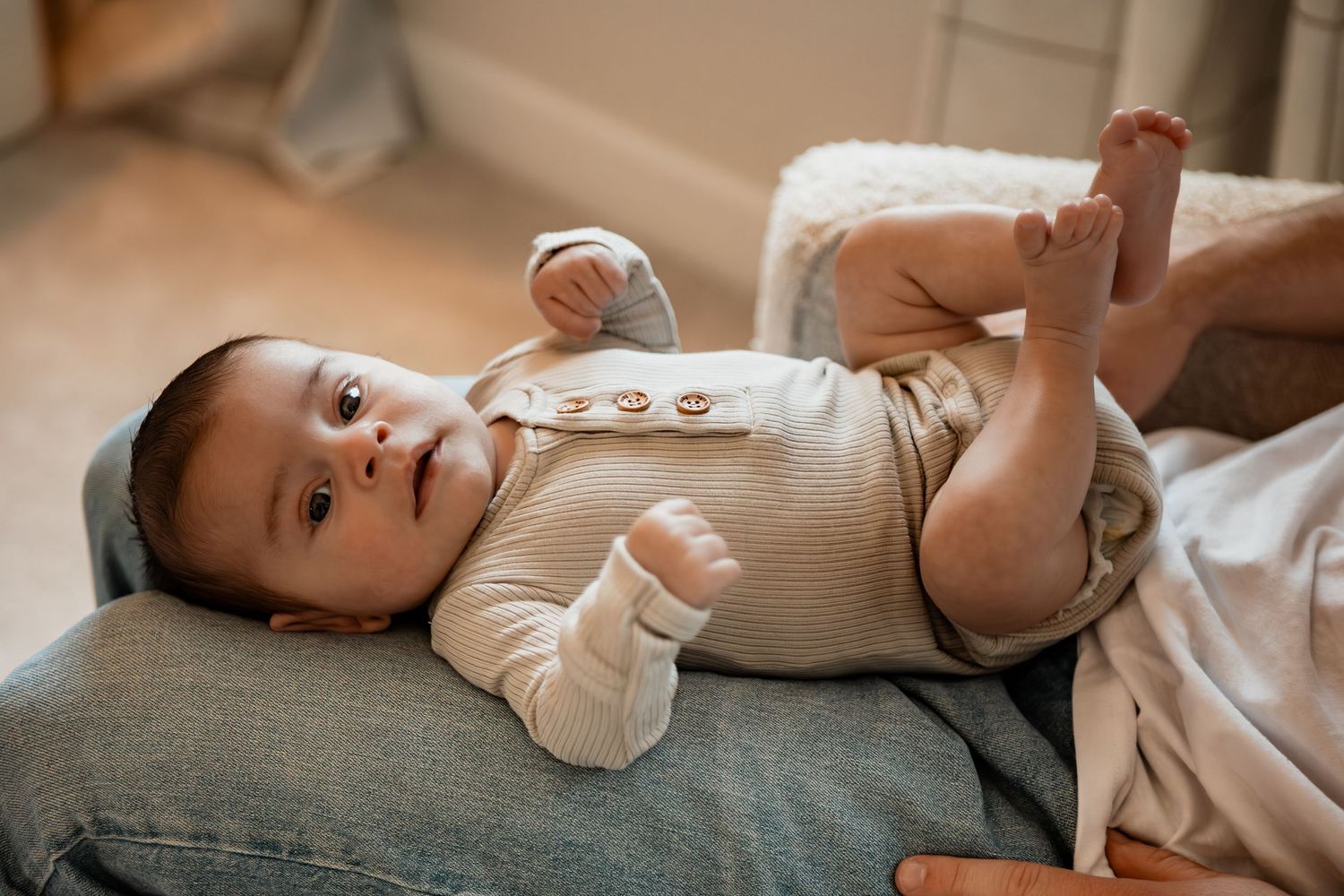 A small baby wearing a white onesie lies peacefully on a gray cushion while looking upward.
