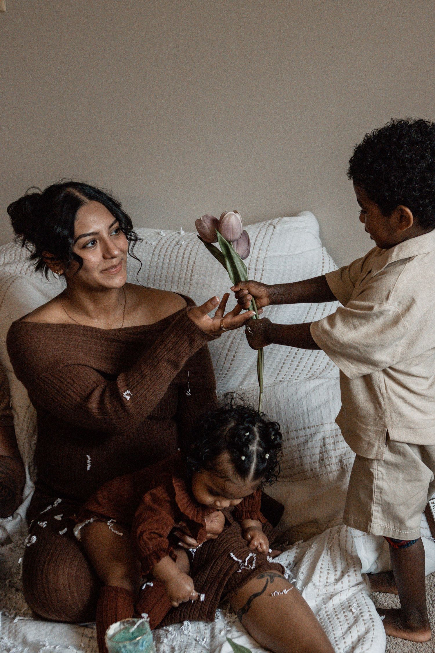 A tender family moment captured on a white bed as a child presents a pink rose in a cozy bedroom setting.