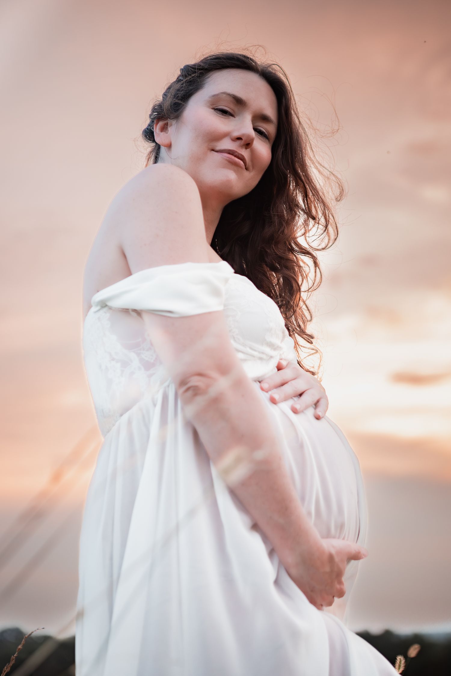A maternity portrait shows a dreamy moment in a white dress against a glowing sunset sky.