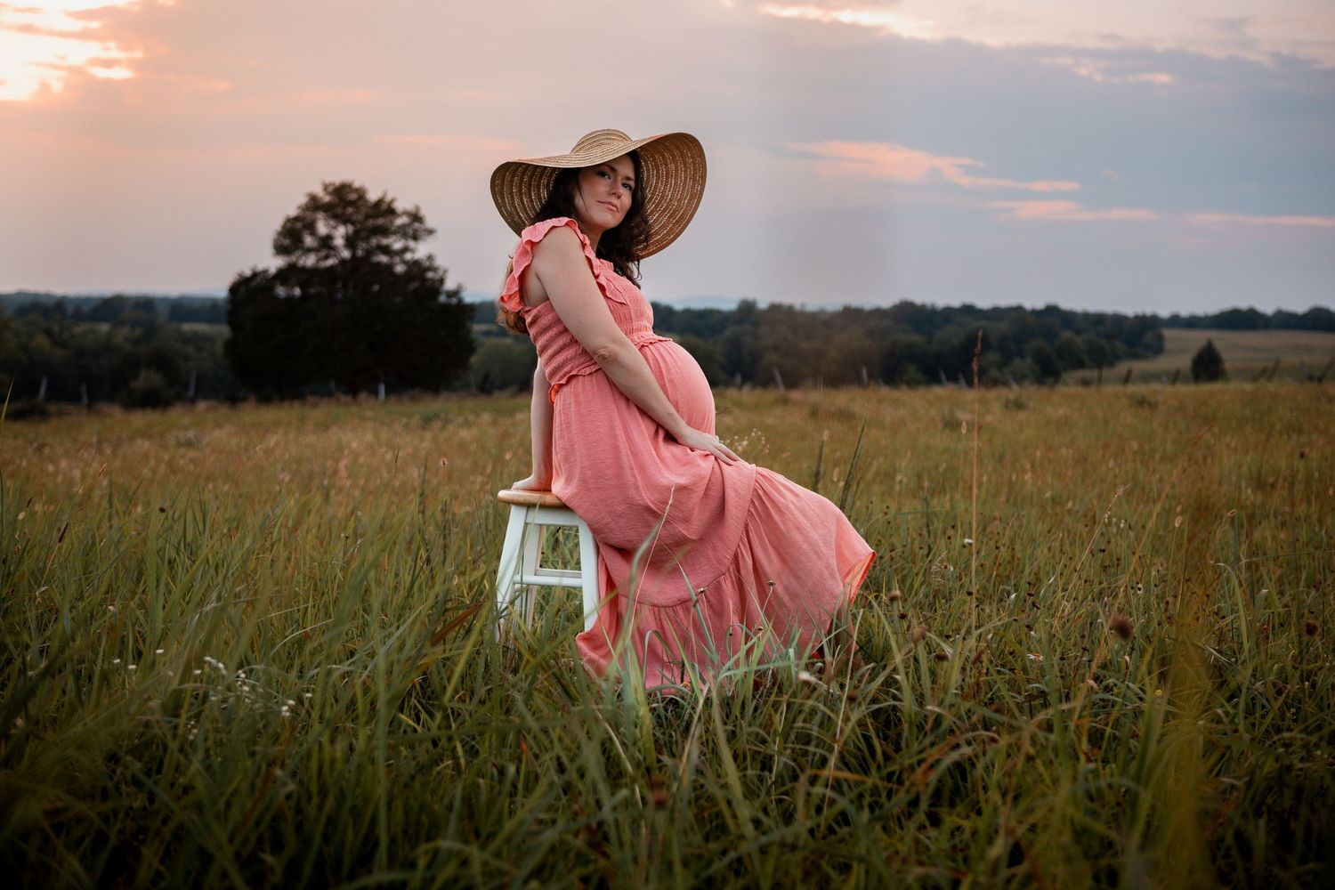 A figure in a flowing pink dress and wide-brimmed hat stands on a chair in a golden field at sunset.