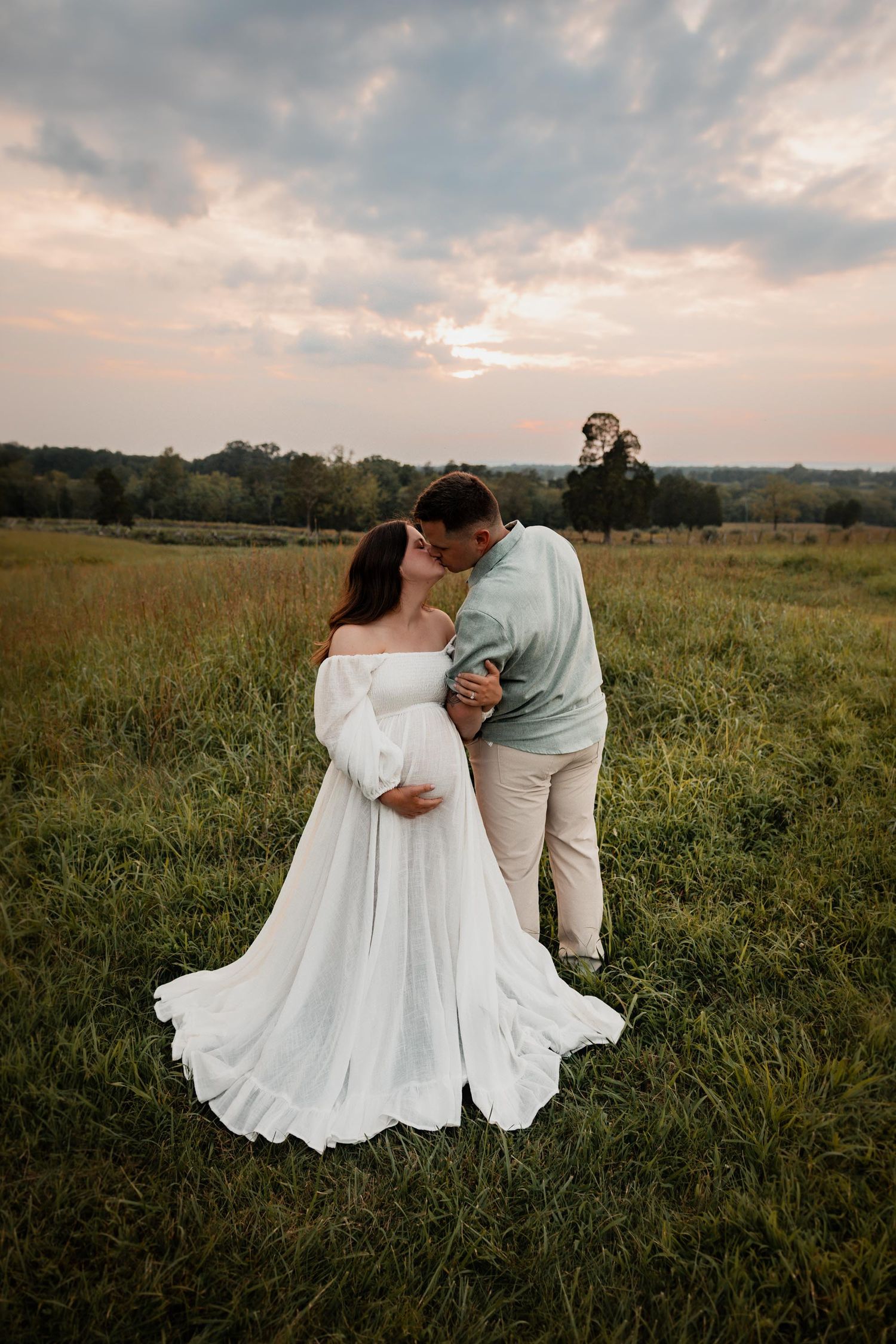 Couple kissing in a field at sunset, woman in white maternity gown, man in light blue shirt, lush green meadow backdrop.