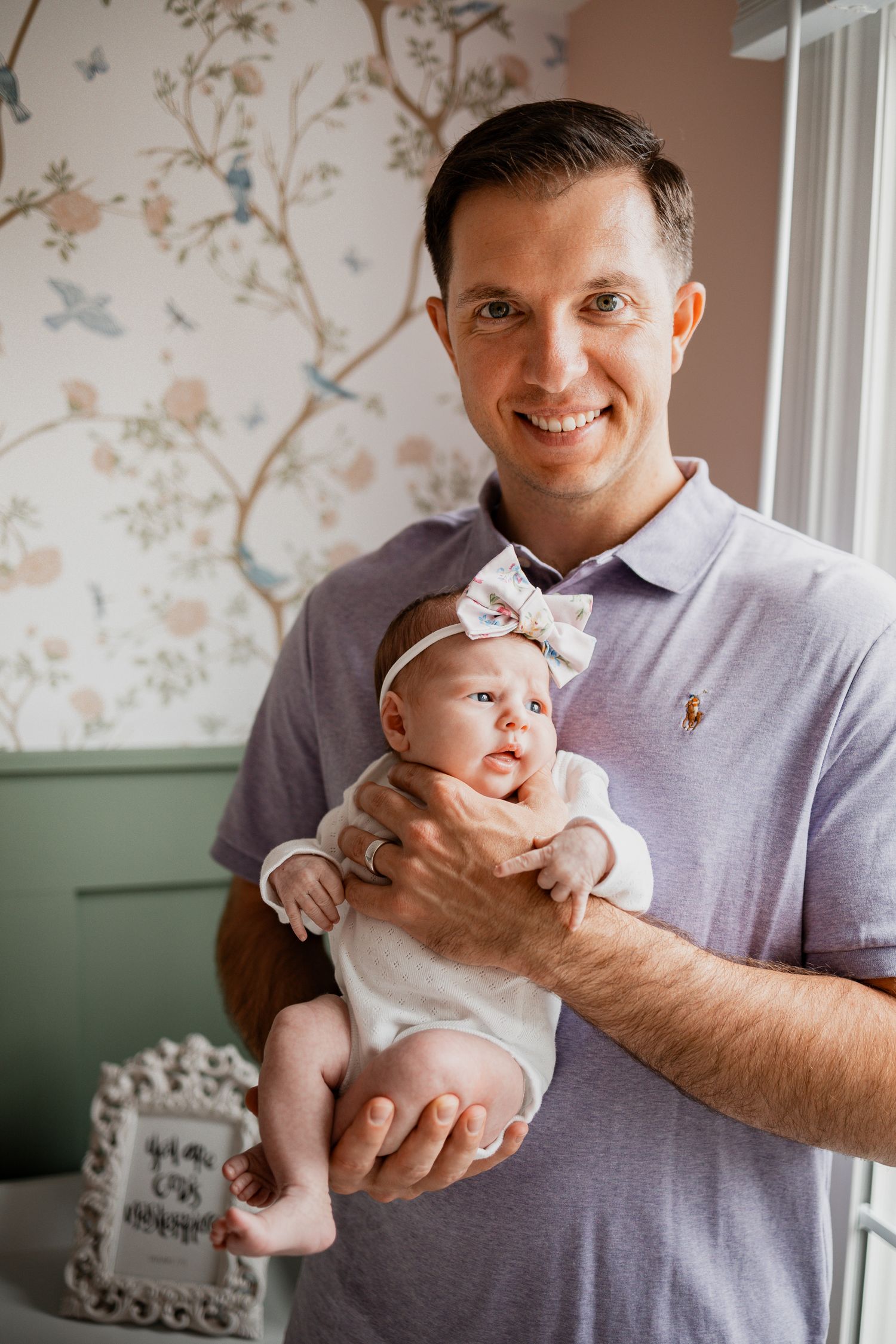 A baby wearing white clothing and bow is held lovingly in a room with floral wallpaper.