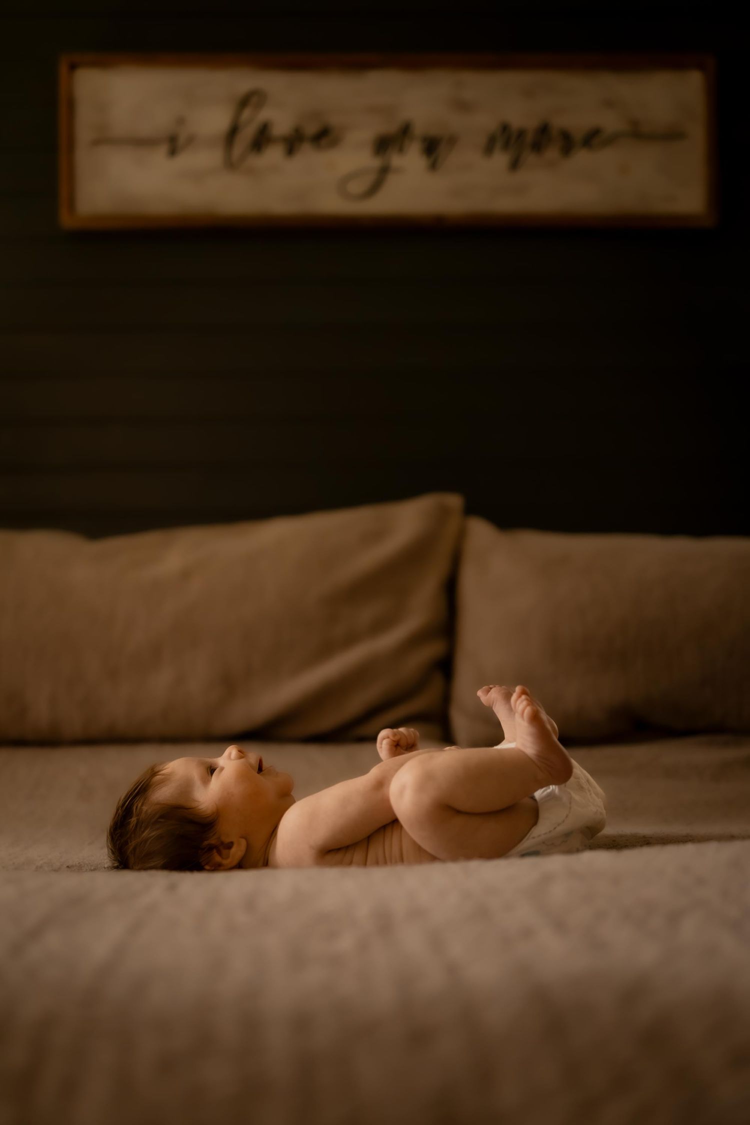 Newborn baby lying on a bed in a diaper beneath an 'I Love You More' wooden sign in soft warm light.