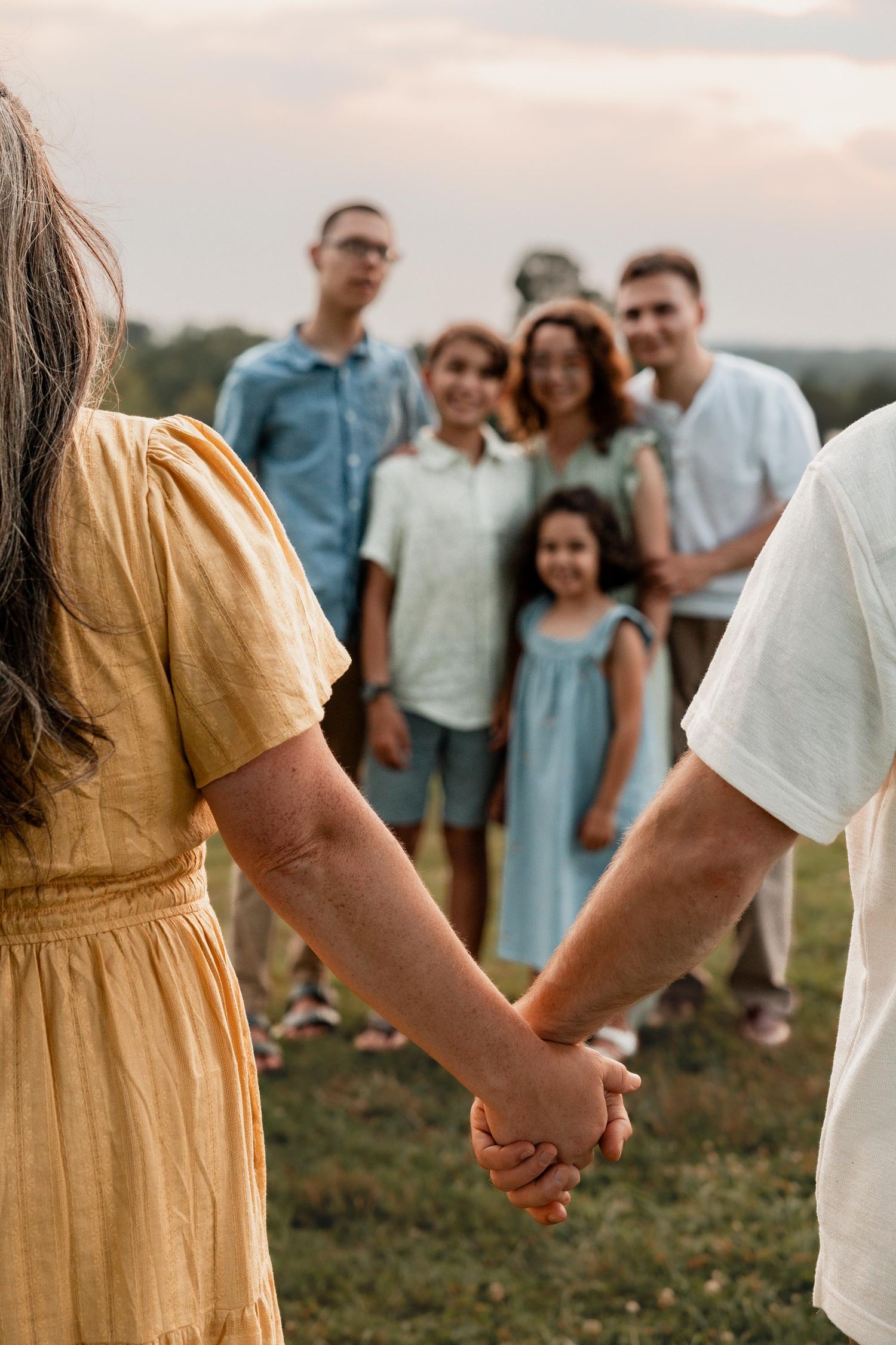 A couple holds hands in a field while a group of smiling family members stands together in the background.