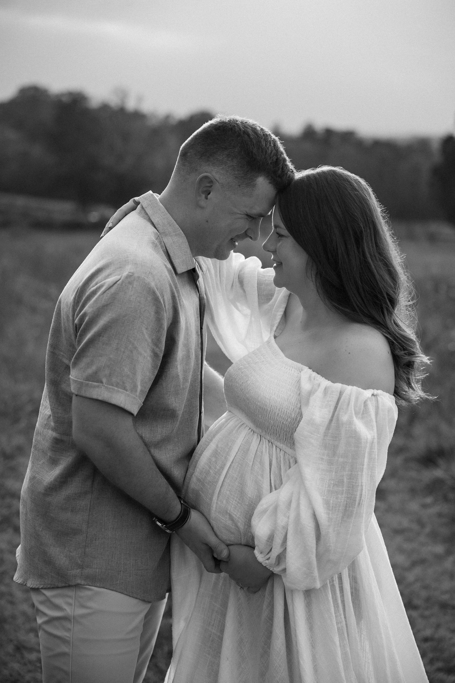 Black and white photo of a smiling couple embracing in a field, woman in white off-shoulder dress, maternity shoot.
