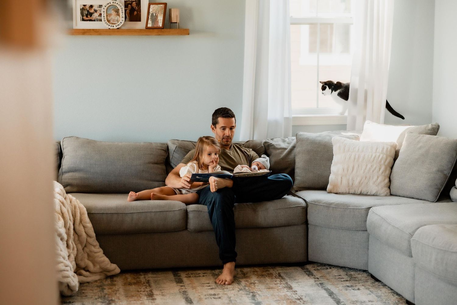 Father and young daughter sitting together on gray sectional sofa reading a book in a cozy living room.