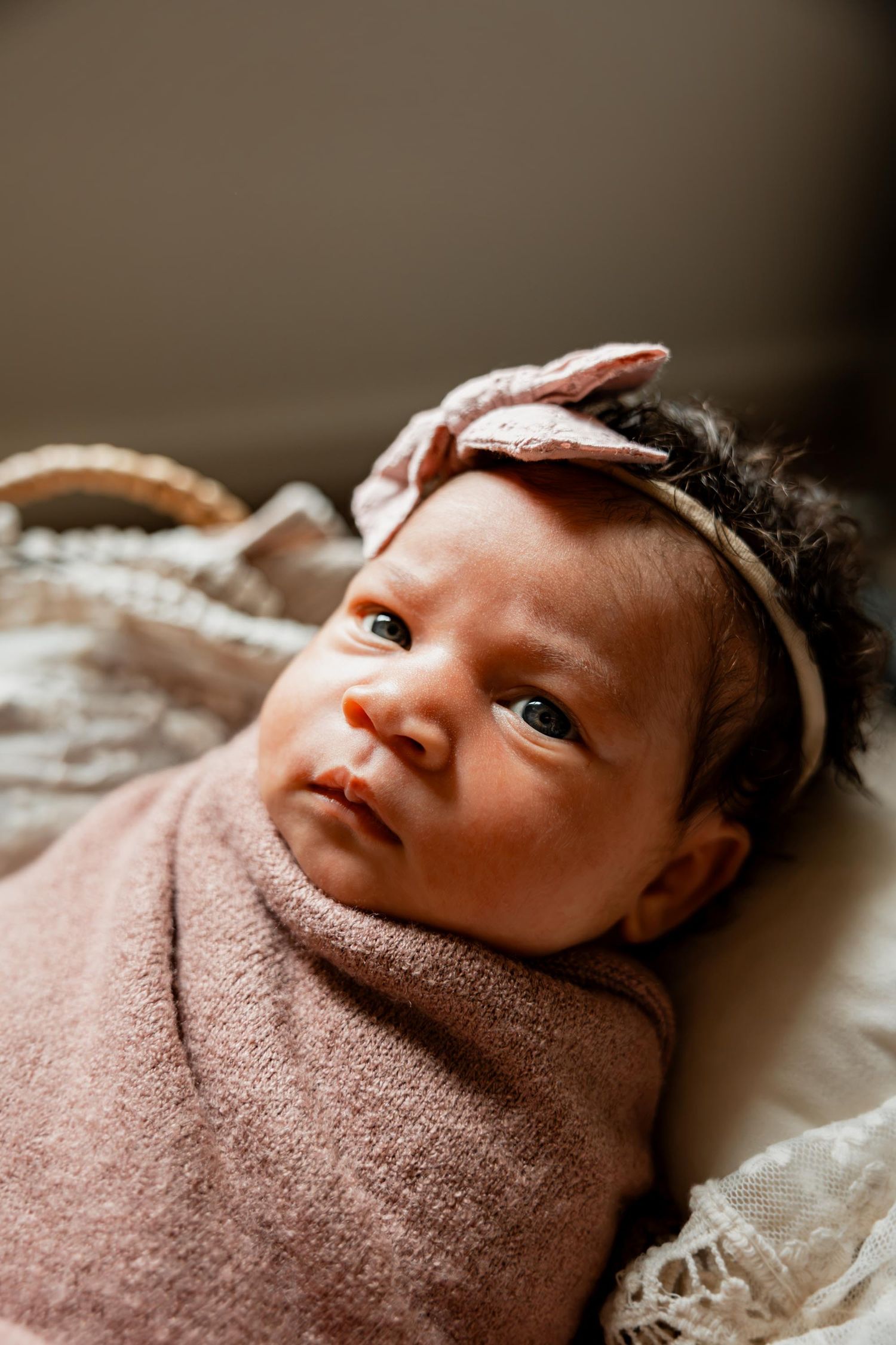 Awake newborn baby girl swaddled in pink wrap with bow headband lying in a wicker basket looking up.