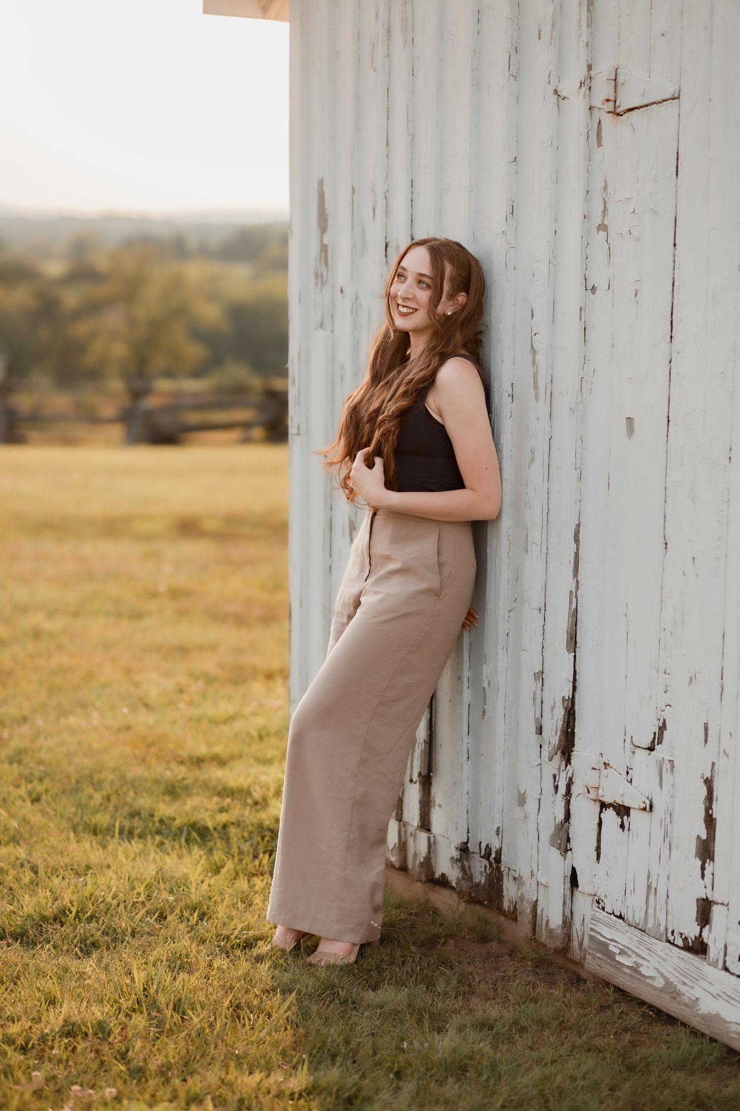 Young woman in beige wide-leg pants and black top leaning against a rustic white barn door in a sunny field.