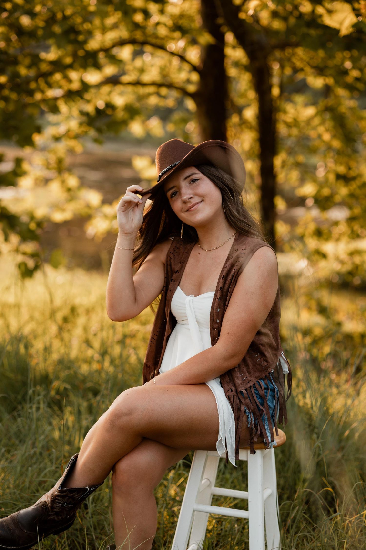 Young woman in white dress, brown vest, cowboy hat, and boots sits on stool outdoors in golden sunlight.