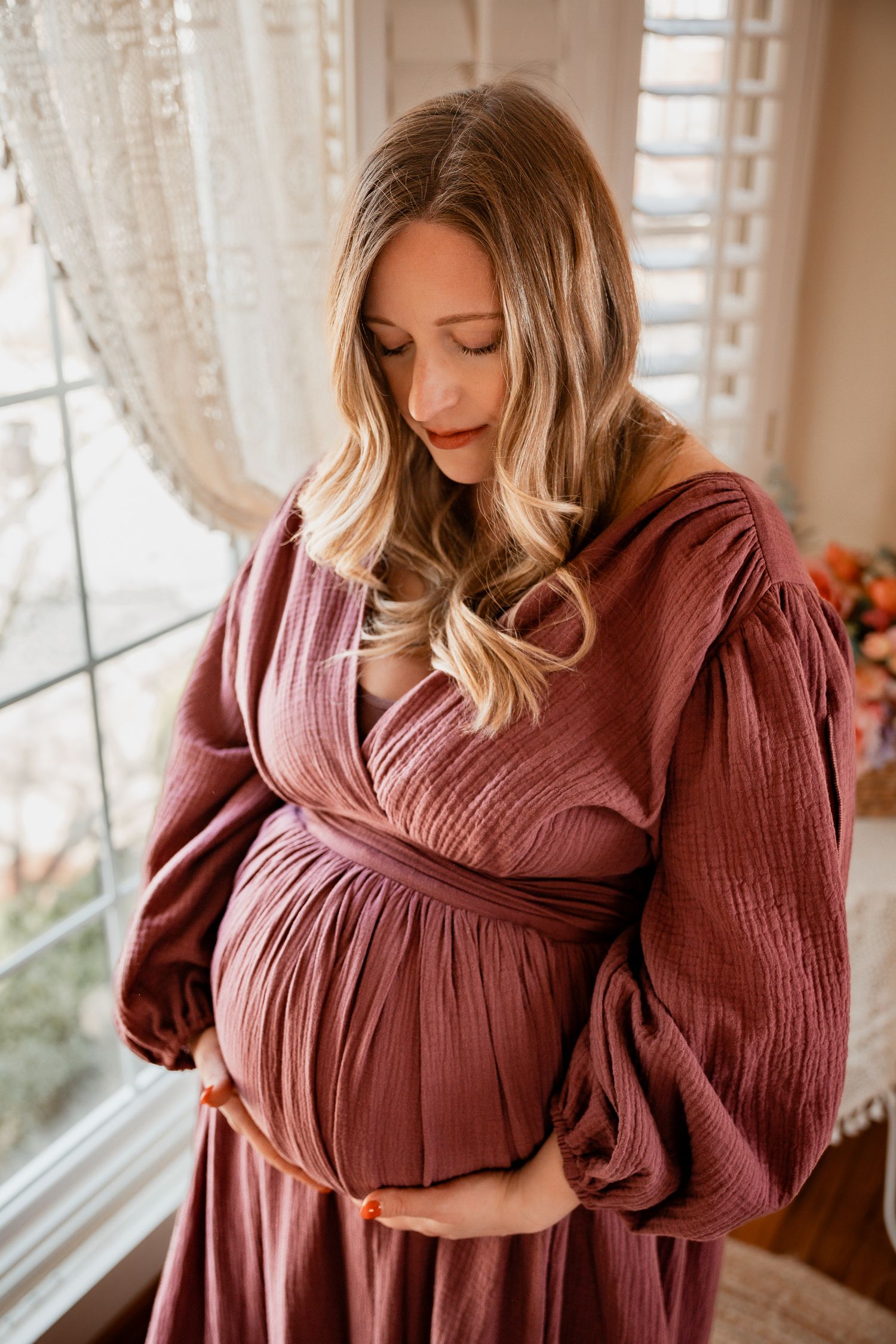 Pregnant woman in mauve dress cradling baby bump by window during maternity photo session.