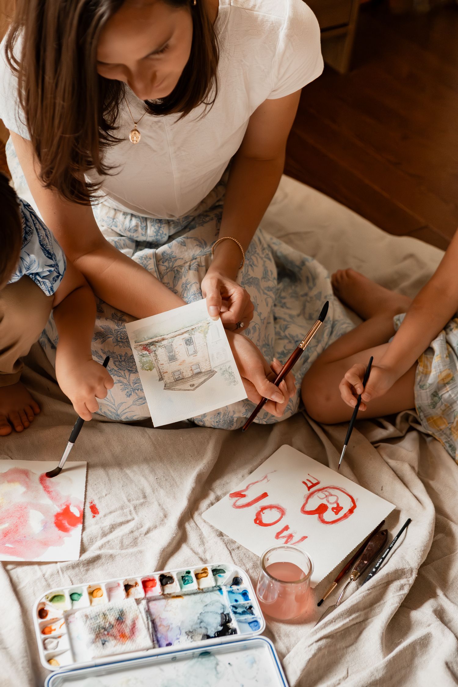 Artist sitting on floor with watercolor supplies painting while wearing a floral dress.