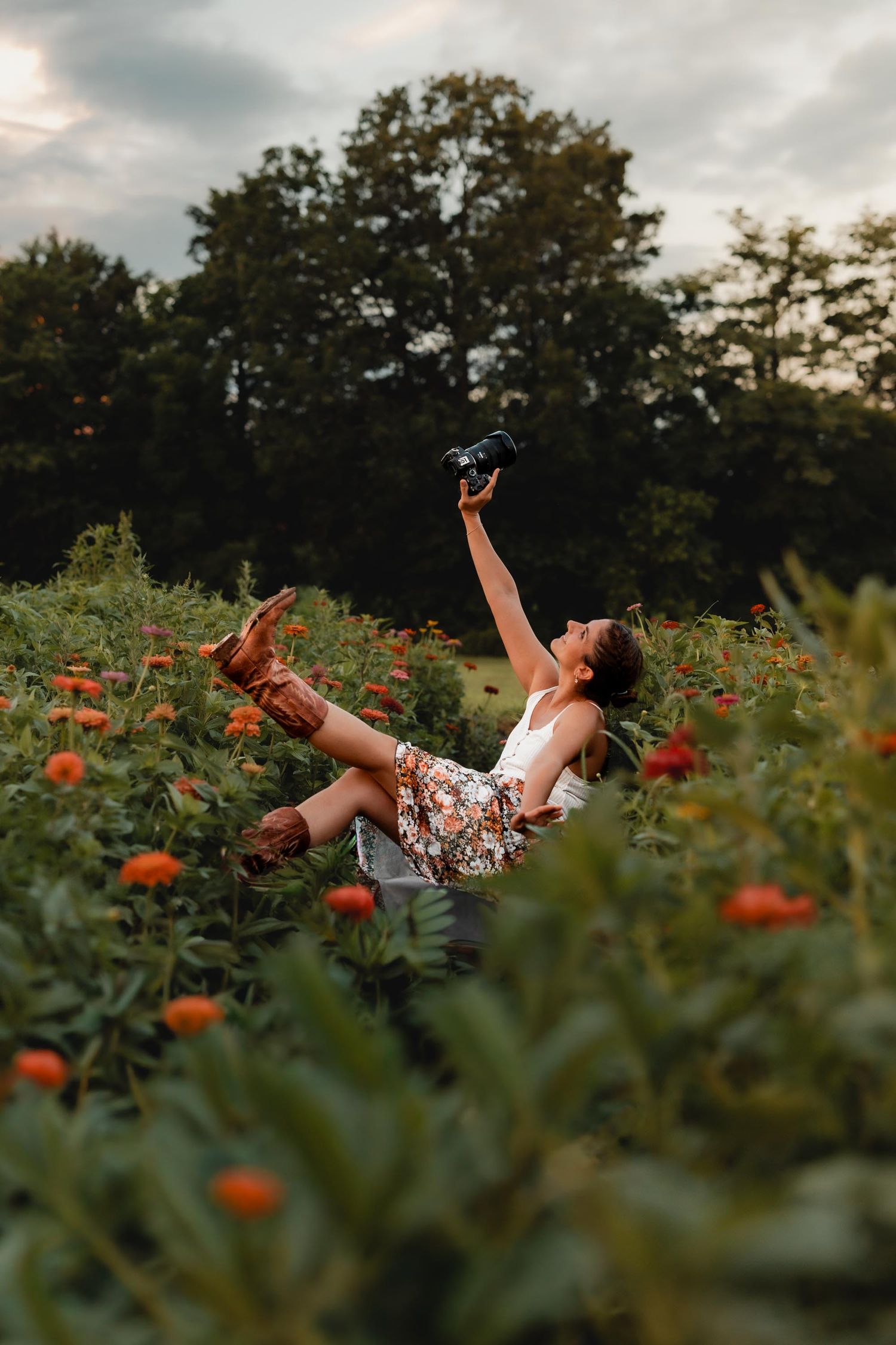 Woman joyfully sitting in a flower field, holding a camera up high, wearing a floral skirt and brown boots.