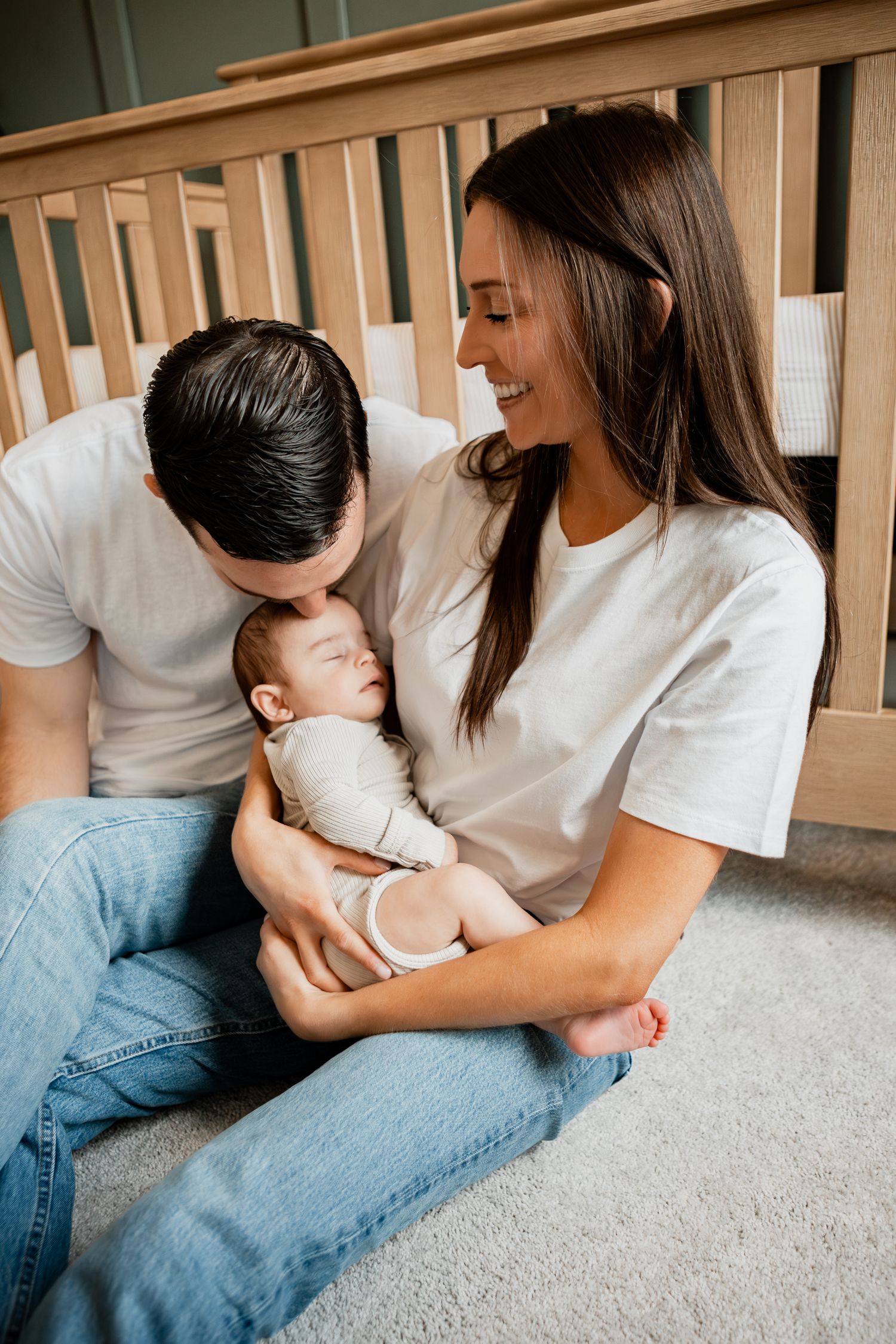 Family bonding moment on carpeted floor wearing matching white t-shirts and blue jeans near a wooden crib.