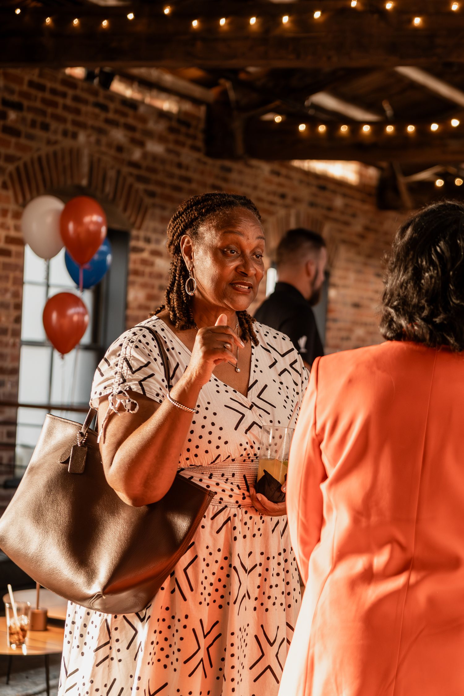 Guests mingling at a party with string lights and orange balloons in a brick-walled venue.