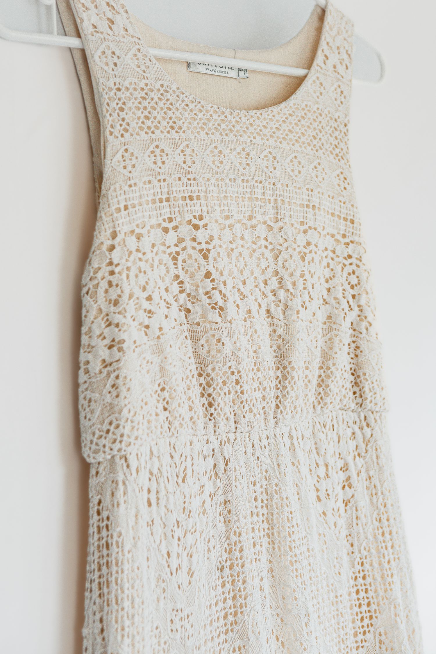 A delicate white lace summer dress with intricate crochet detailing hangs on a hanger against a light background.