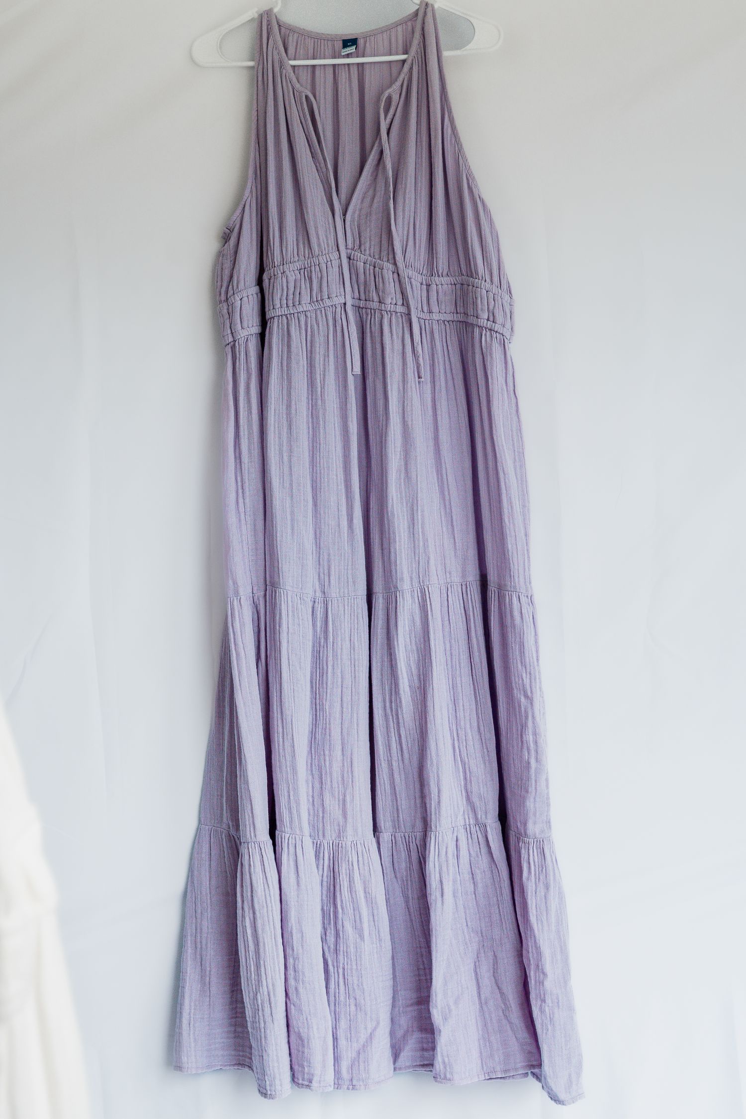 A flowy lavender tiered maxi dress with V-neck design and sleeveless cut hangs on a white hanger against a light wall.