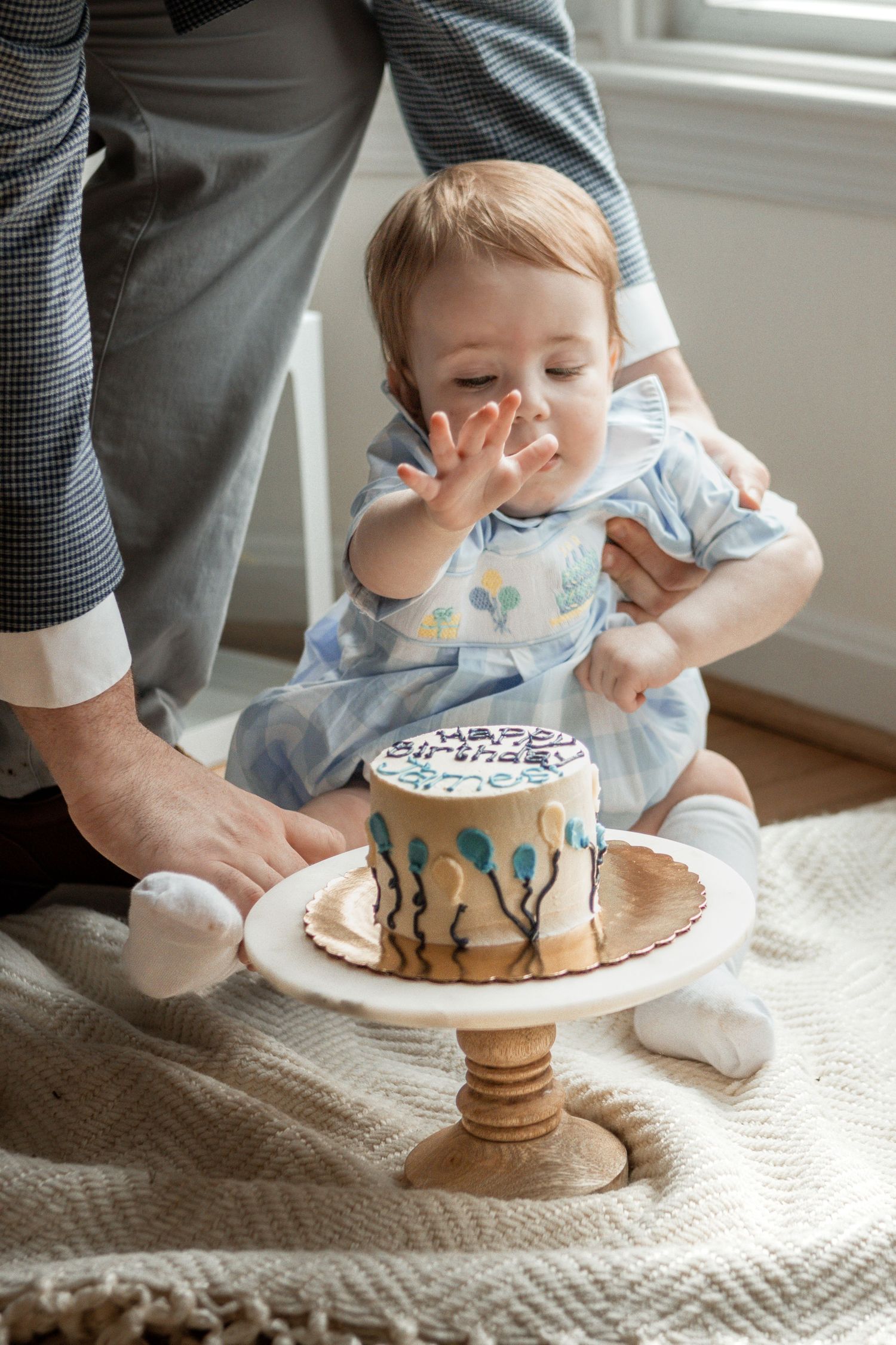 Small child in blue romper reaches toward a decorated birthday cake on wooden cake stand while sitting on textured blanket.